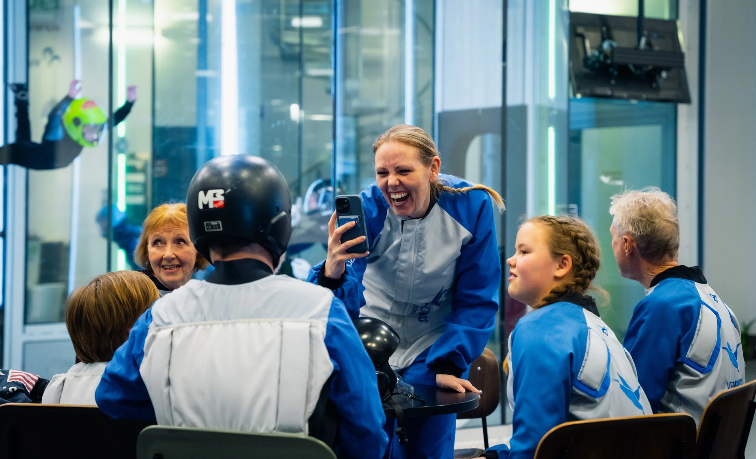 En gruppe mennesker iført blå og hvite jakker, med en kvinne i midten som ler og tar et bilde med telefonen, mens resten sitter rundt bordet, med en bakgrunn av en vindtunnel