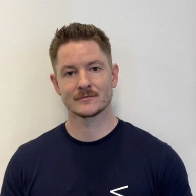 Portrettbilde av en mann med kort, lyst hår og skjegg, iført en mørk T-skjorte, stående mot en hvit vegg.