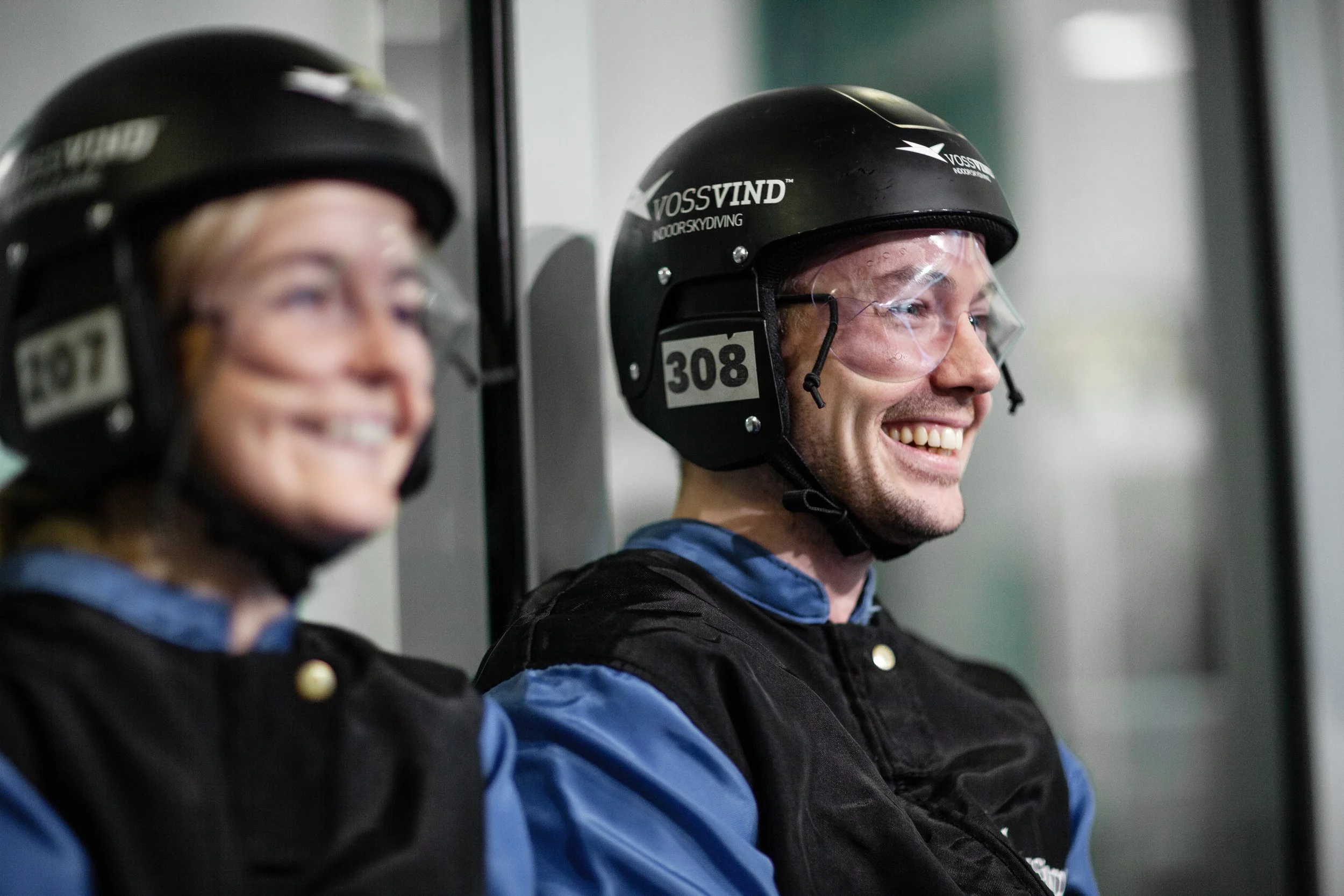 To personer iført hjelmer og beskyttelsesutstyr, sitter ved vindtunnellen, ventende, smilende.
