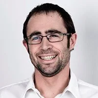 Portrett av en smilende mann med briller og mørkt hår, iført hvit skjorte, mot en lys bakgrunn.