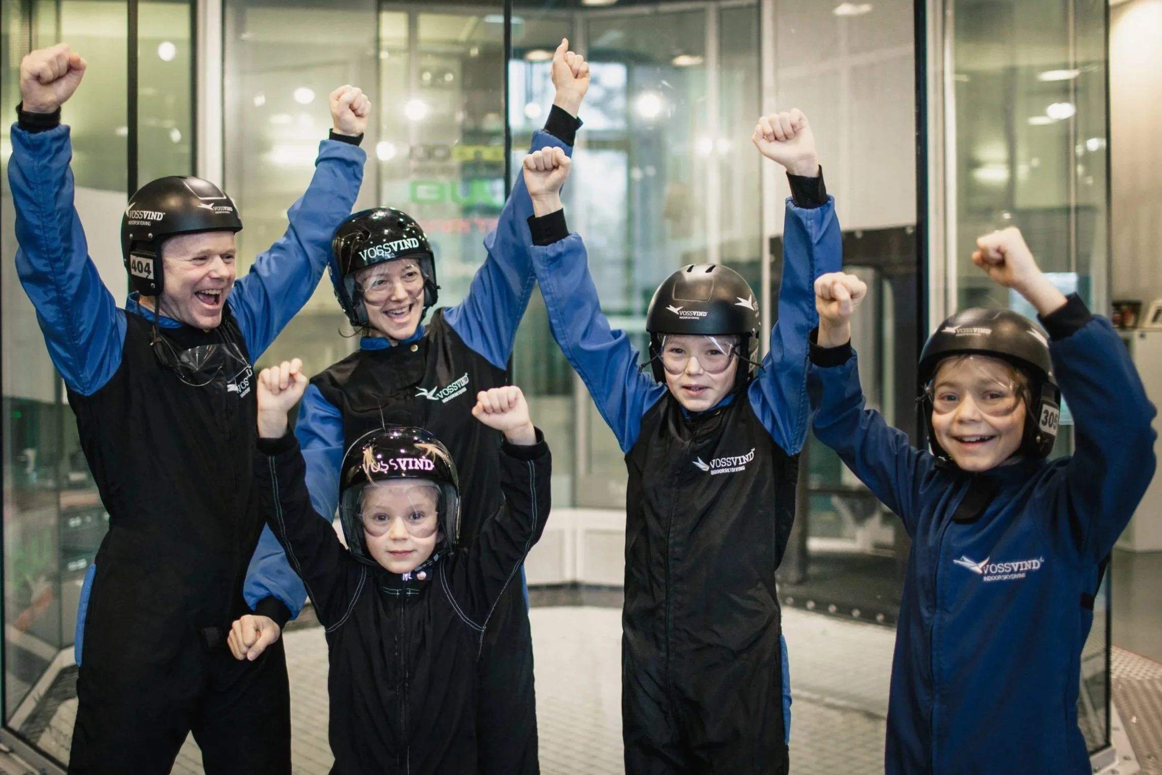 Fem personer i skydiverbekledning feirer med armene hevet innendørs, alle smiler og har på hjelm og jakker med logo.