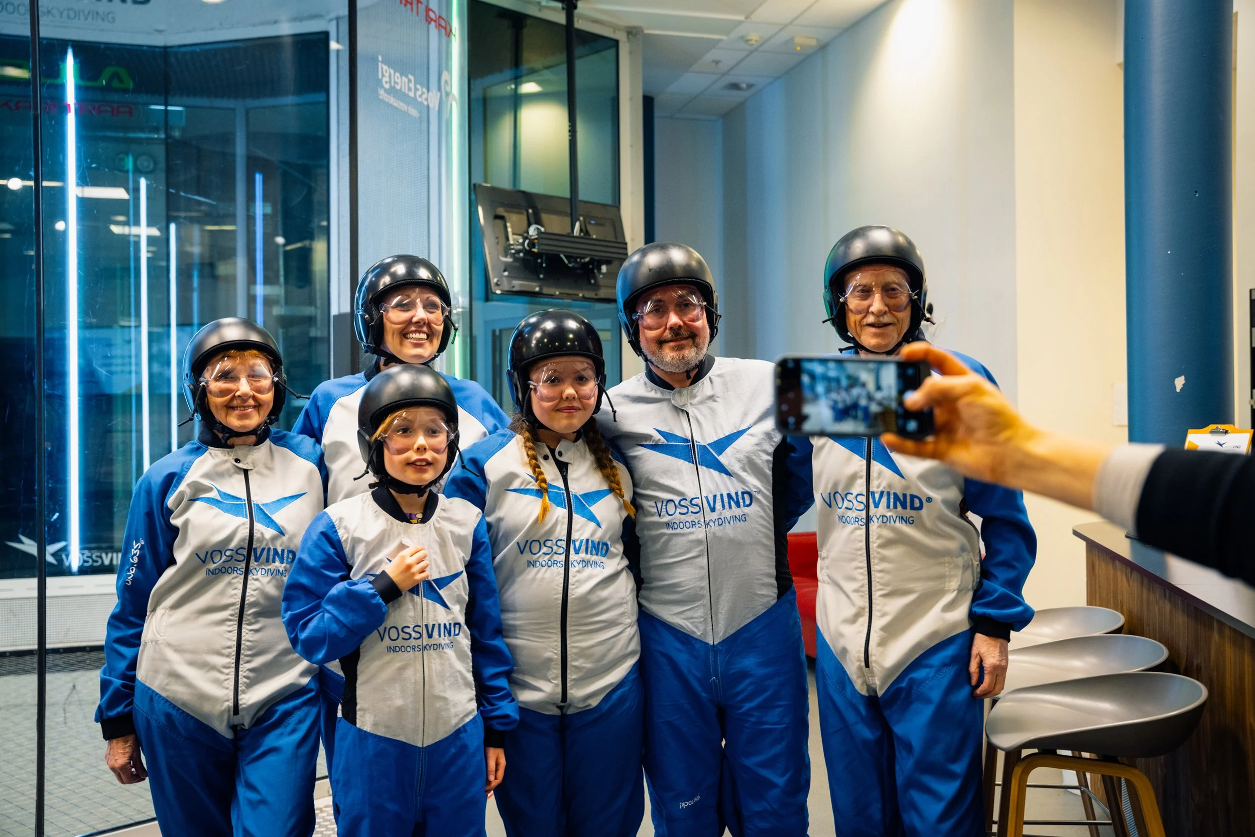 En gruppe personer i ulik alder i dress med logoen Voss Vind Indoor Skydiving tar bildet, alle med hjelm og vernebriller, innendørs skydiving fasilitet.