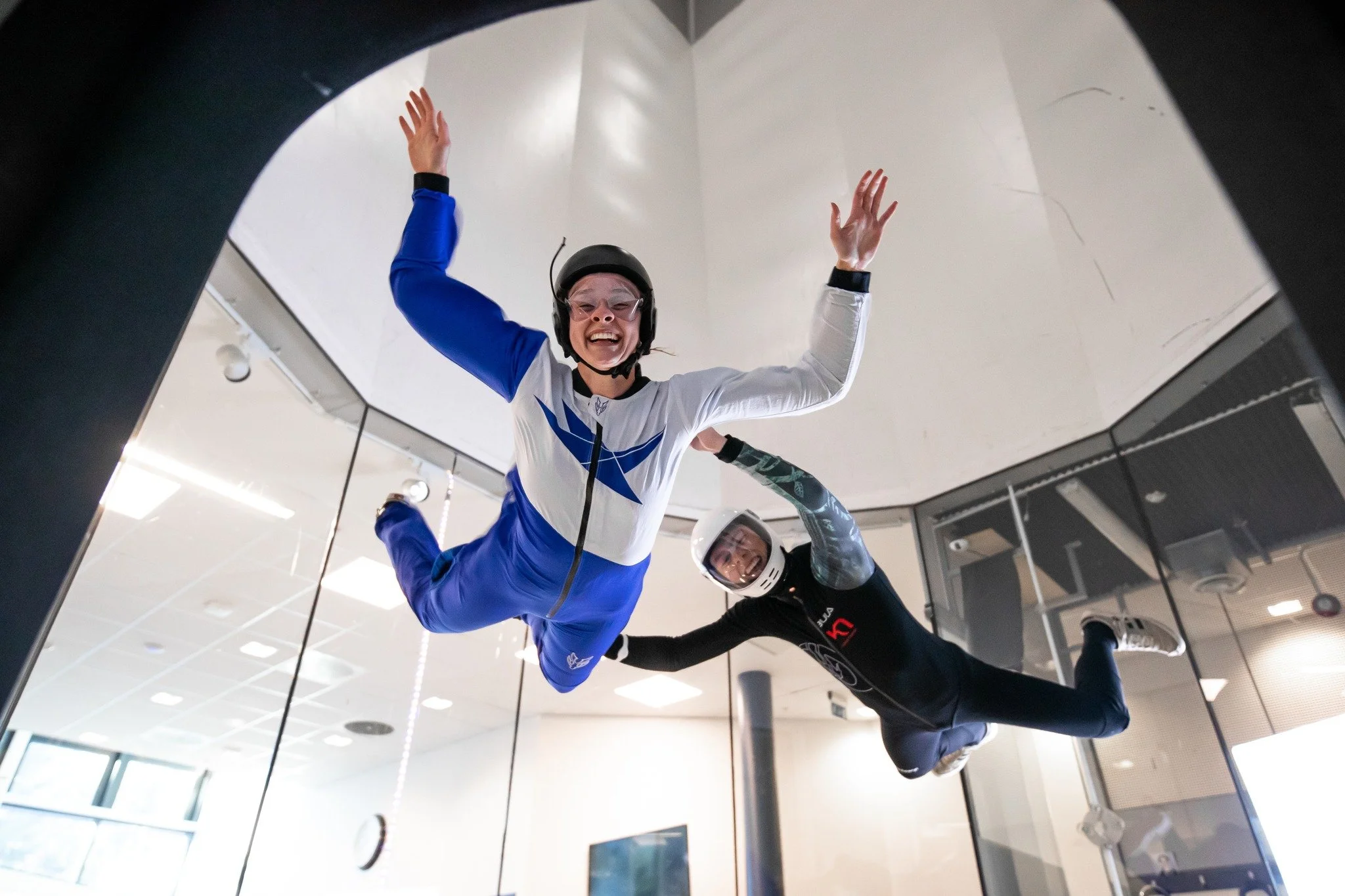 To personer flyvende i vindtunnel innendørs, begge iført hjelmer og utstyr, med en person foran og den andre bak, begge har utstrakte armer og glade ansikter. Instruktør og elev.