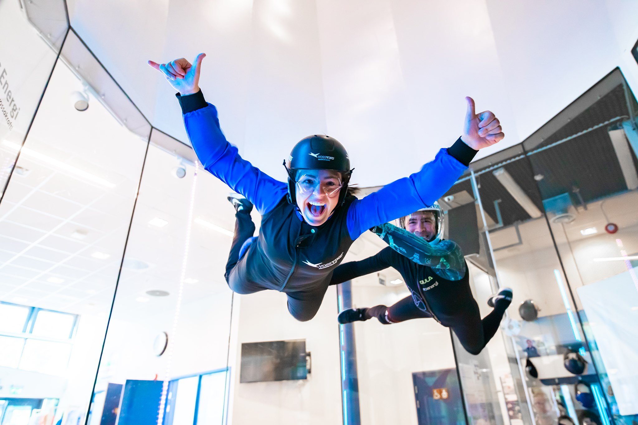 To personer flyvende i vindtunnel innendørs, begge iført hjelmer og utstyr, med en person foran og den andre bak, begge har utstrakte armer og glade ansikter.