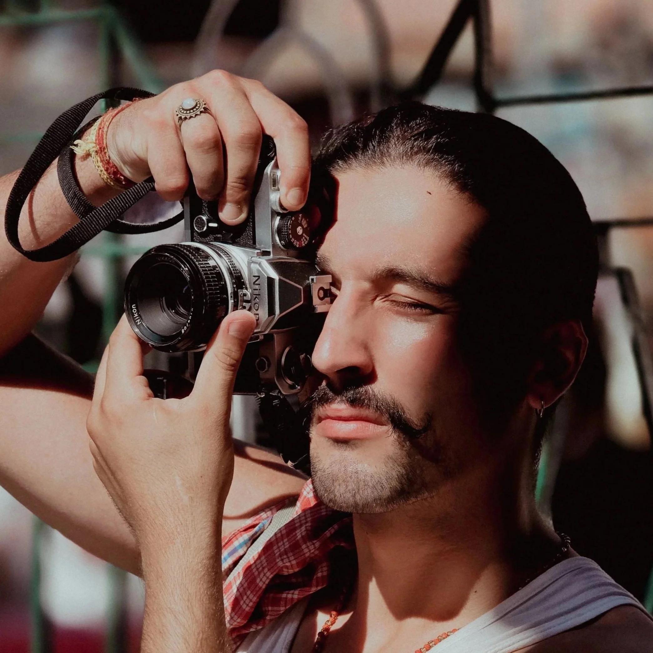 Homme avec une barbe, coiffé en arrière, ferme les yeux, tenant un appareil photo Nikon 35mm, portant un t-shirt gris et quelques bijoux.