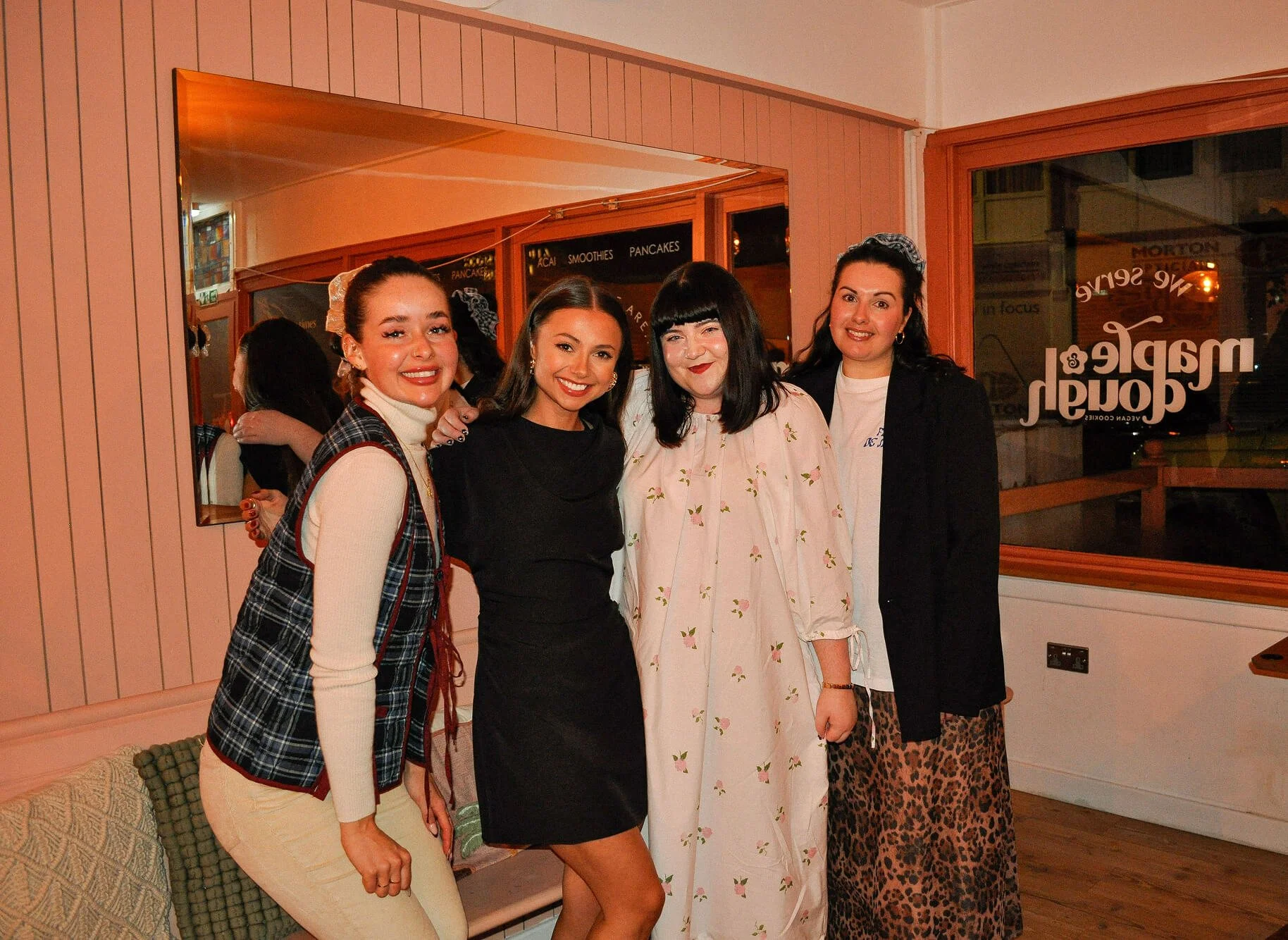 Four women standing together inside a restaurant, smiling at the camera, with pink walls and mirror behind them.