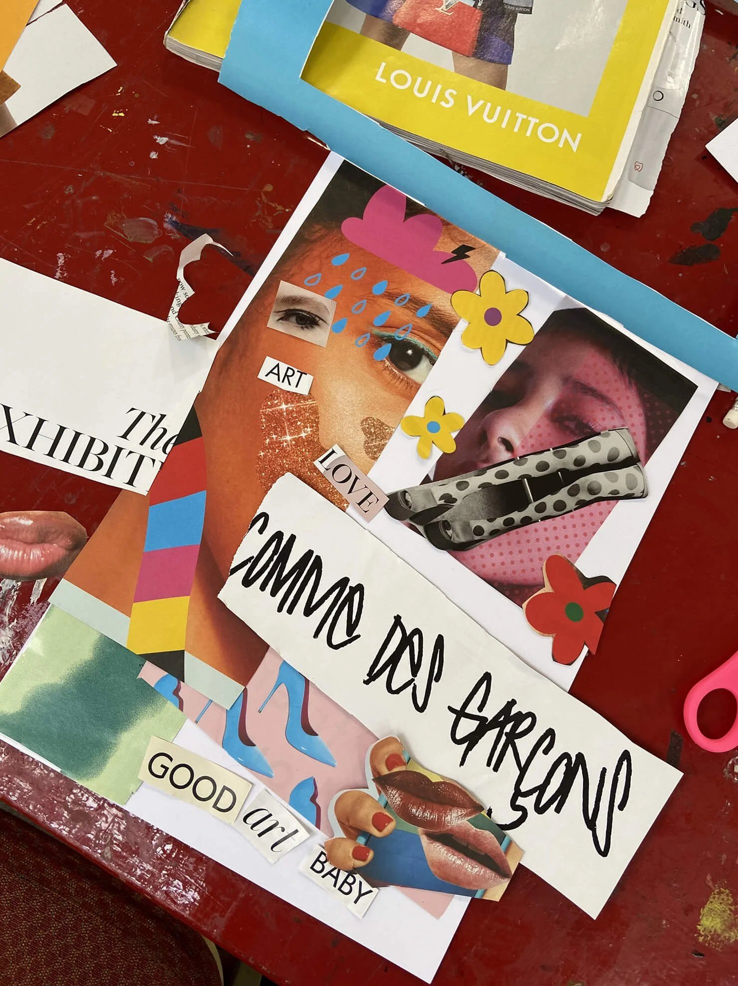 A collage art project on a table featuring images of faces, lips, rain, and flowers with cut-out words like 'ART,' 'LOVE,' and 'COMMON FORGIVENESS.' There are also decorative elements like a polka-dot strap and scissors, with a Louis Vuitton magazine or brochure in the background.
