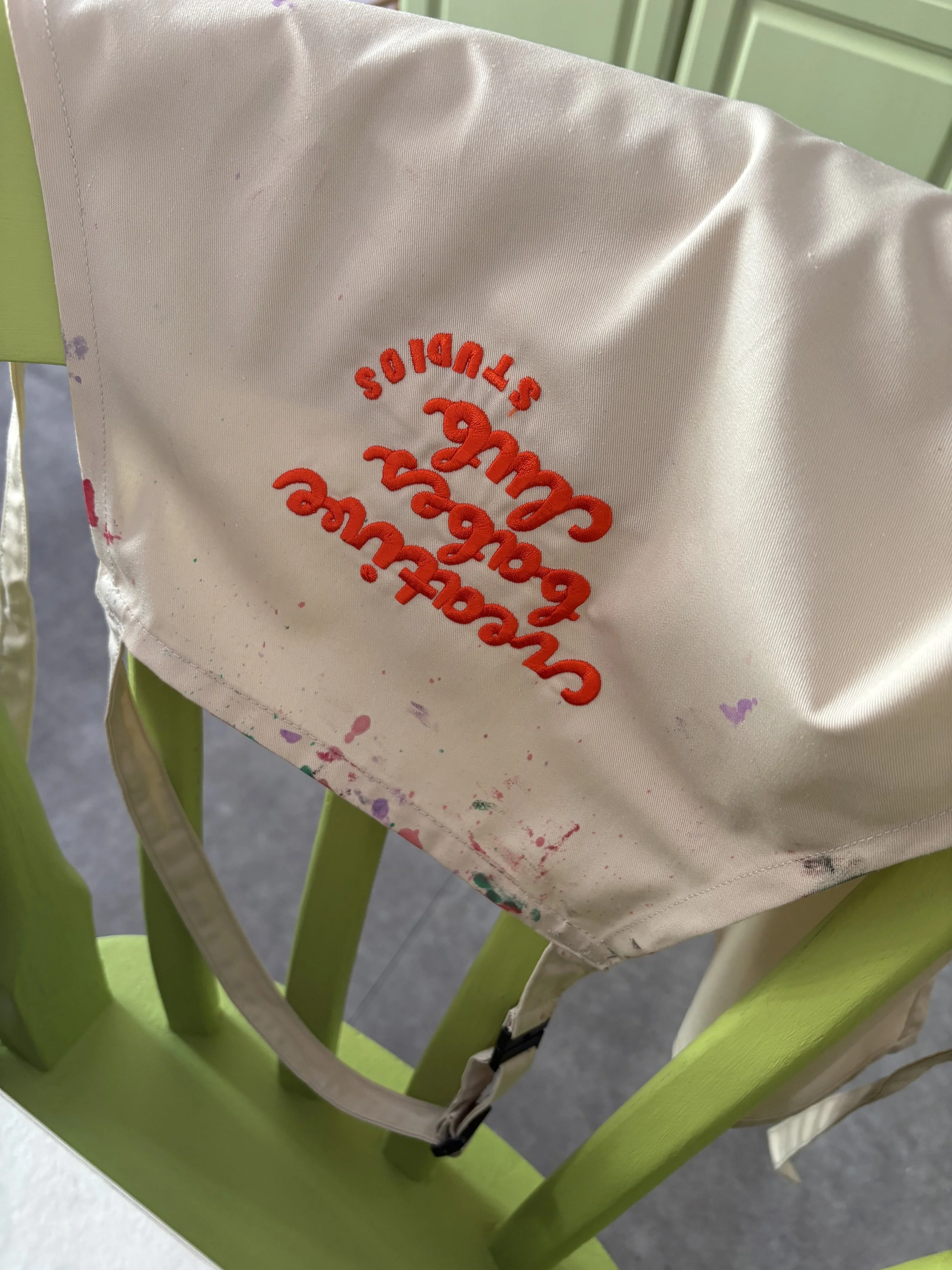 A 'Creative Babes Club Studios' Logo embroidered Apron, covered in colourful paint splatters, is draped over the back of a Lime Green chair.