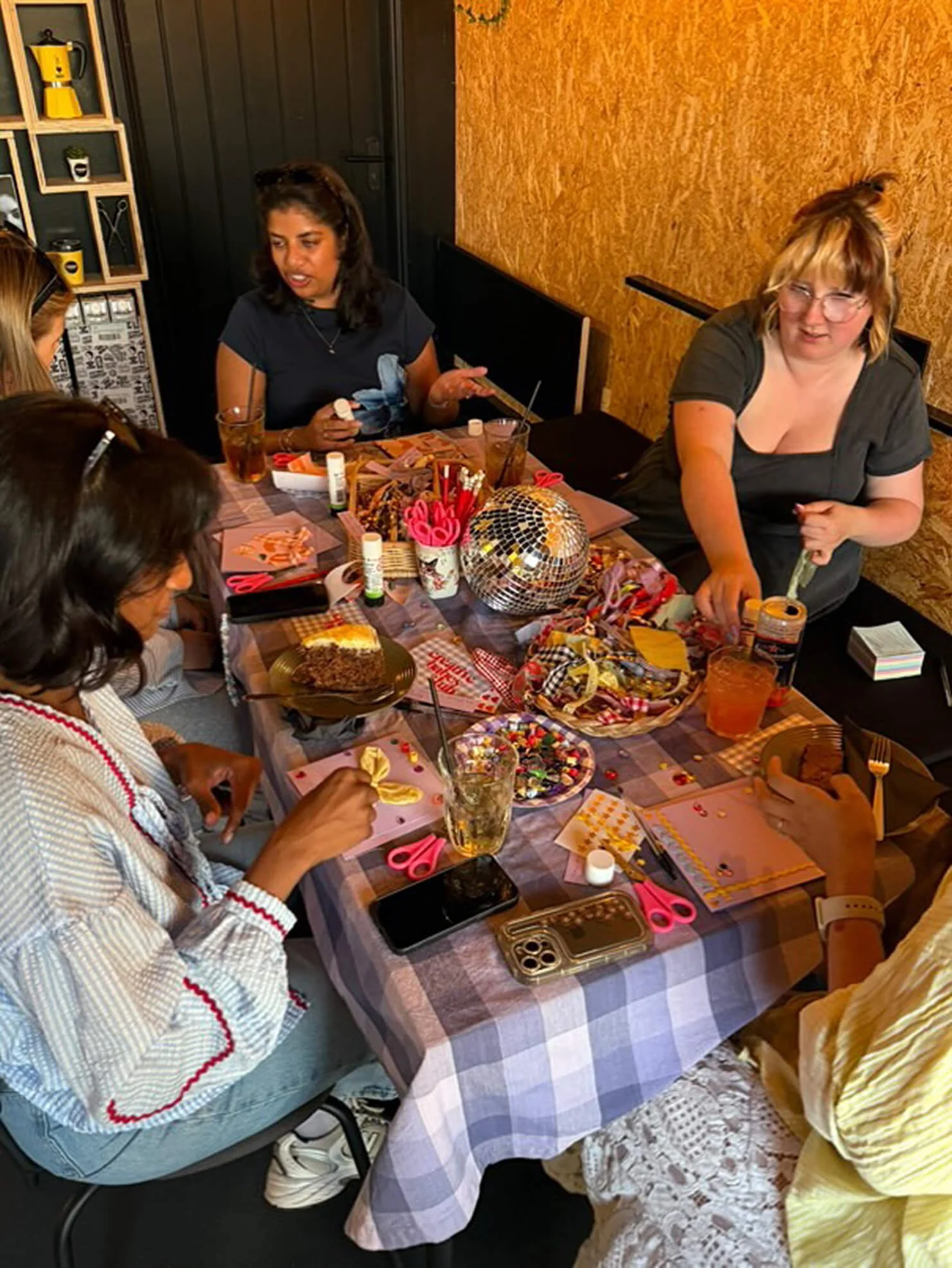A group of women gathered around a table decorated with party supplies, cake, and drinks, celebrating together in a cozy indoor setting.
