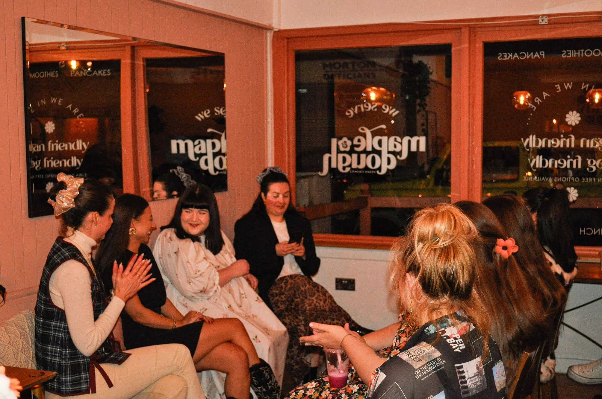 A group of women are sitting together in a cozy indoor space, chatting and laughing. The background includes windows with reflections and signs for a restaurant's offerings.
