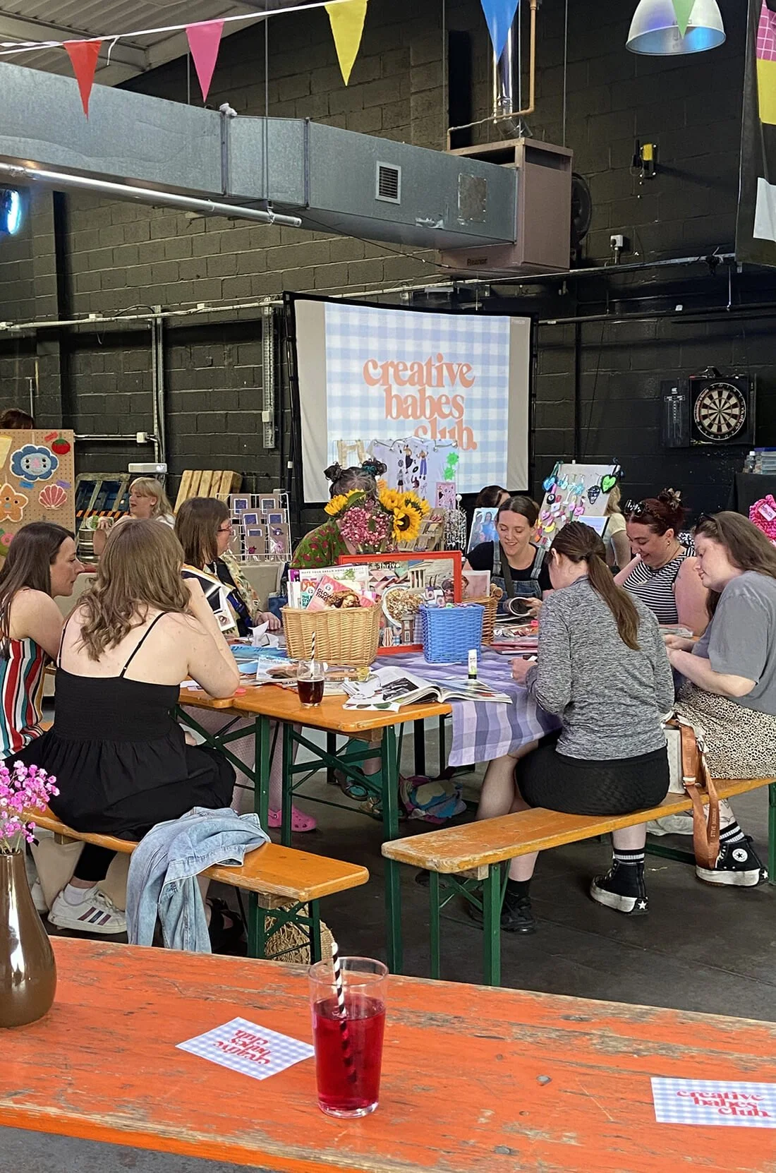 A group of women sitting around a table at a Creative Babes Club event, with decorations, flowers, and displayed crafts, in an industrial-style venue with black walls and colorful bunting.