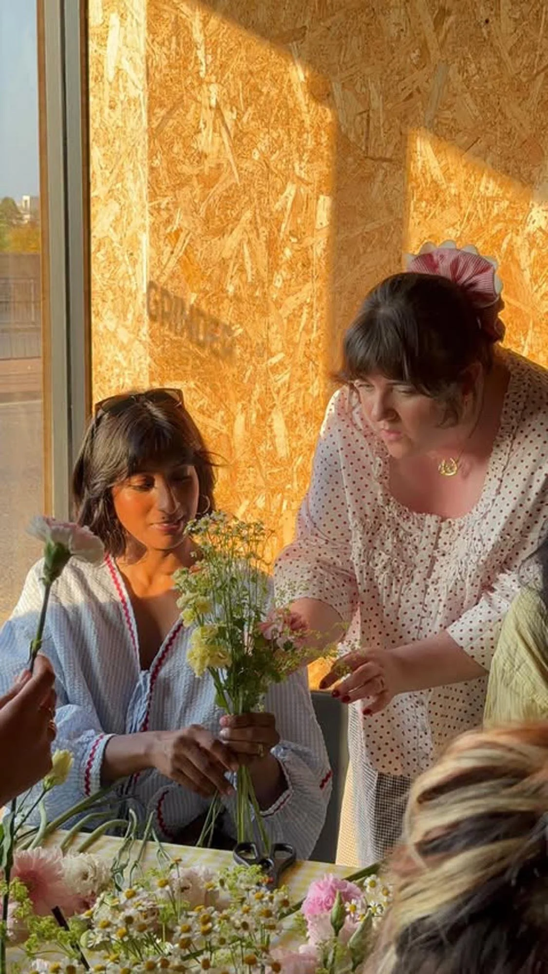 Two women, one seated and the other standing, arranging flowers on a table with various flowers, in a bright room with wood-paneled walls and sunlight coming through a window.