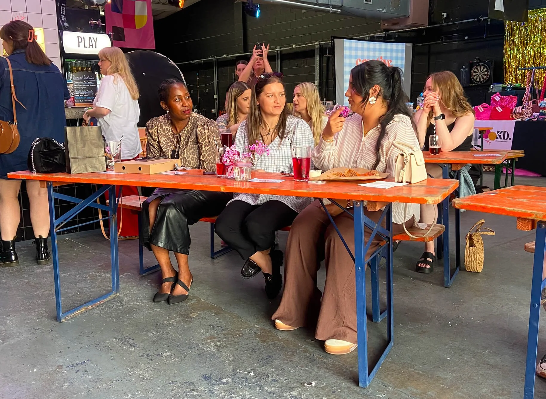 Group of women sitting at orange tables in a bar or restaurant, chatting and drinking, with people standing in the background.