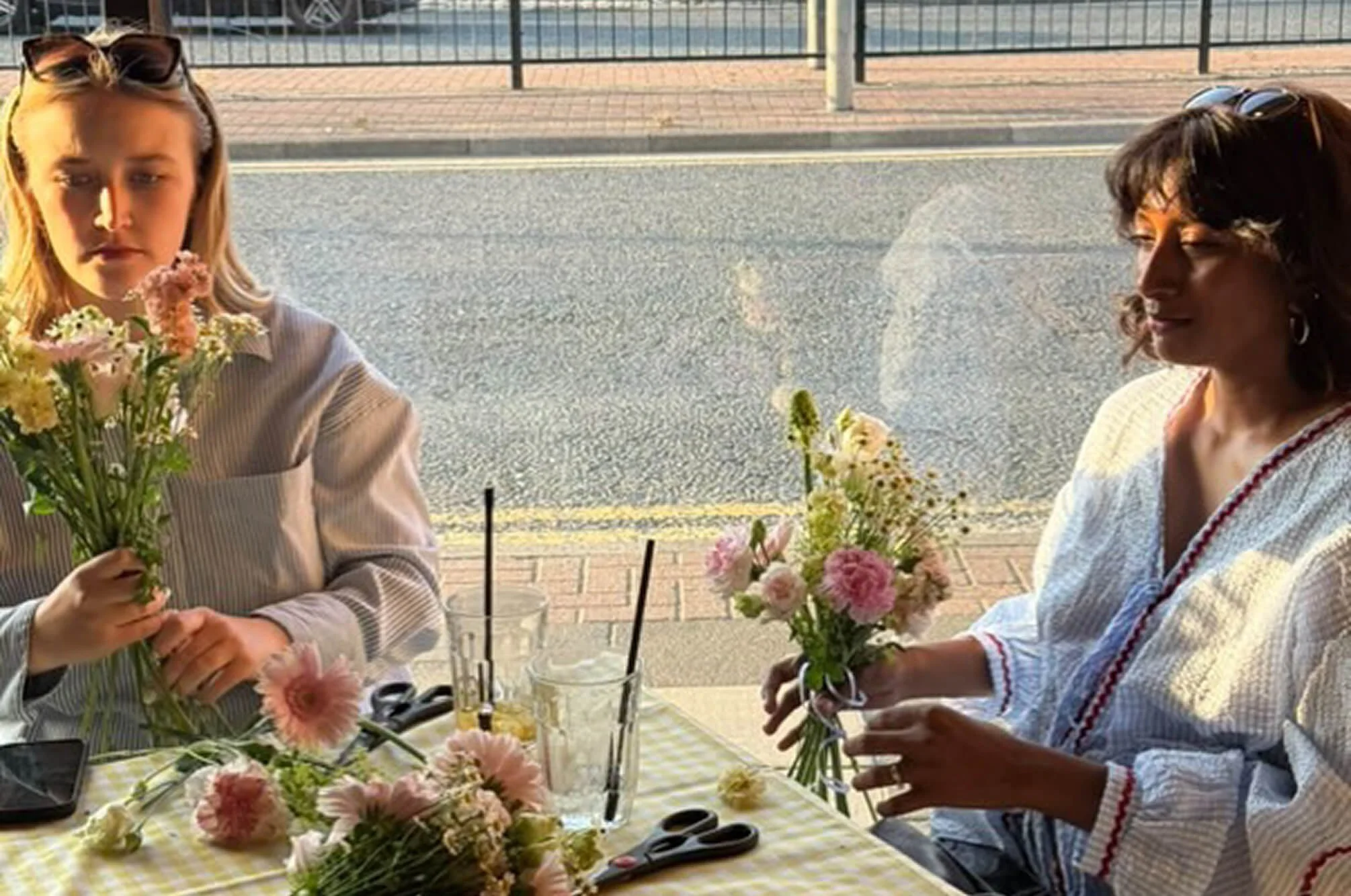 Two women sitting at a table outside, creating flower arrangements with pink, yellow, and white flowers. Sunlight casts shadows on their faces, and on the table are scissors and glasses with drinks.
