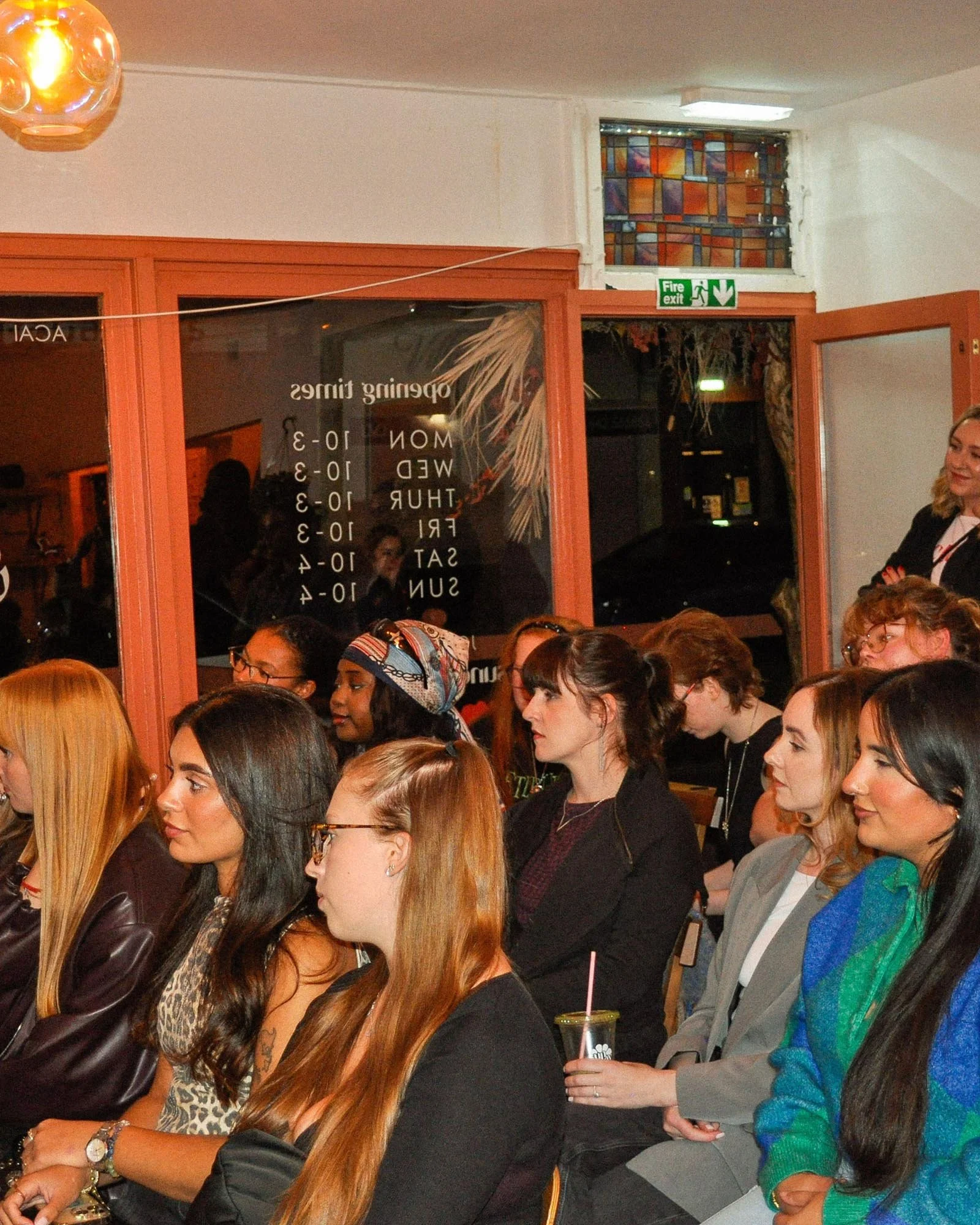 A group of women seated indoors, attentively listening, with some holding drinks, in a casual setting with a large window and a sign listing opening times.