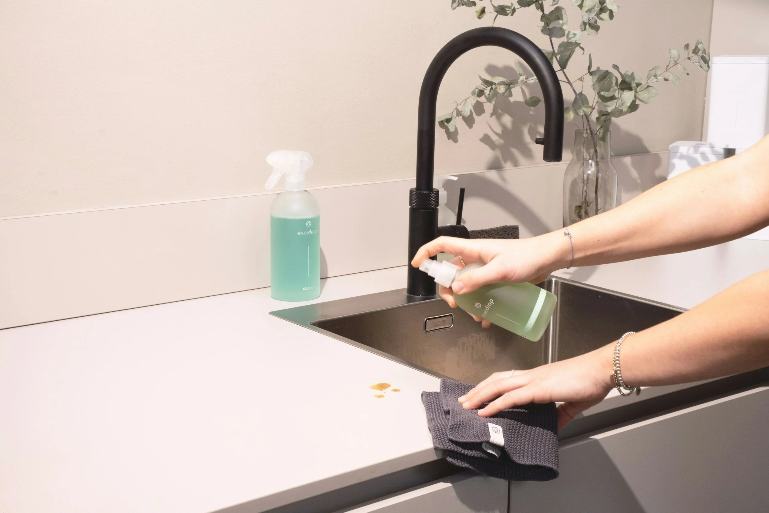 Person spraying green liquid hand sanitizer into a gray towel at a kitchen sink, with a black faucet, a spray bottle on the countertop, and a vase with eucalyptus leaves in the background.