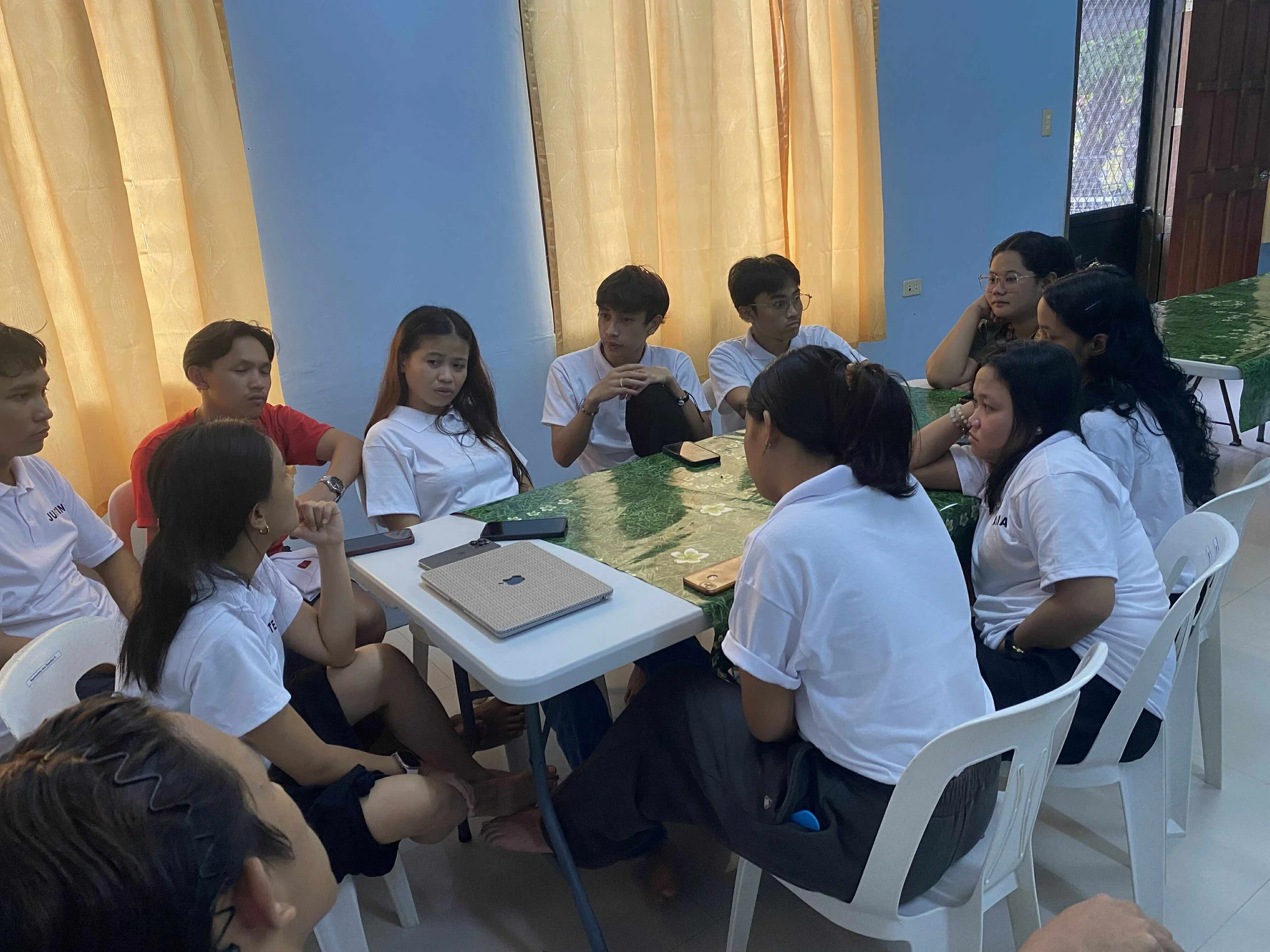 Een groep tieners, enkele vrouwen en mannen, zitten aan een tafel in een kamer met blauwe muren en gele gordijnen, ze lijken een discussie of gesprek te voeren.