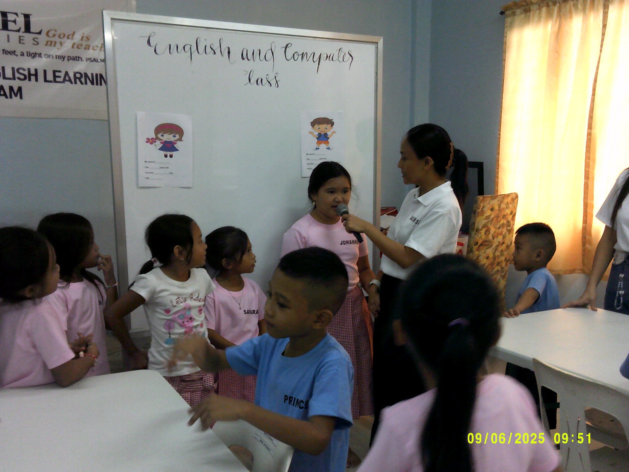 Een klaslokaal met jonge kinderen die deelnemen aan een les Engels. Een vrouw houdt een microfoon voor een meisje dat spreekt, terwijl andere leerlingen toekijken.