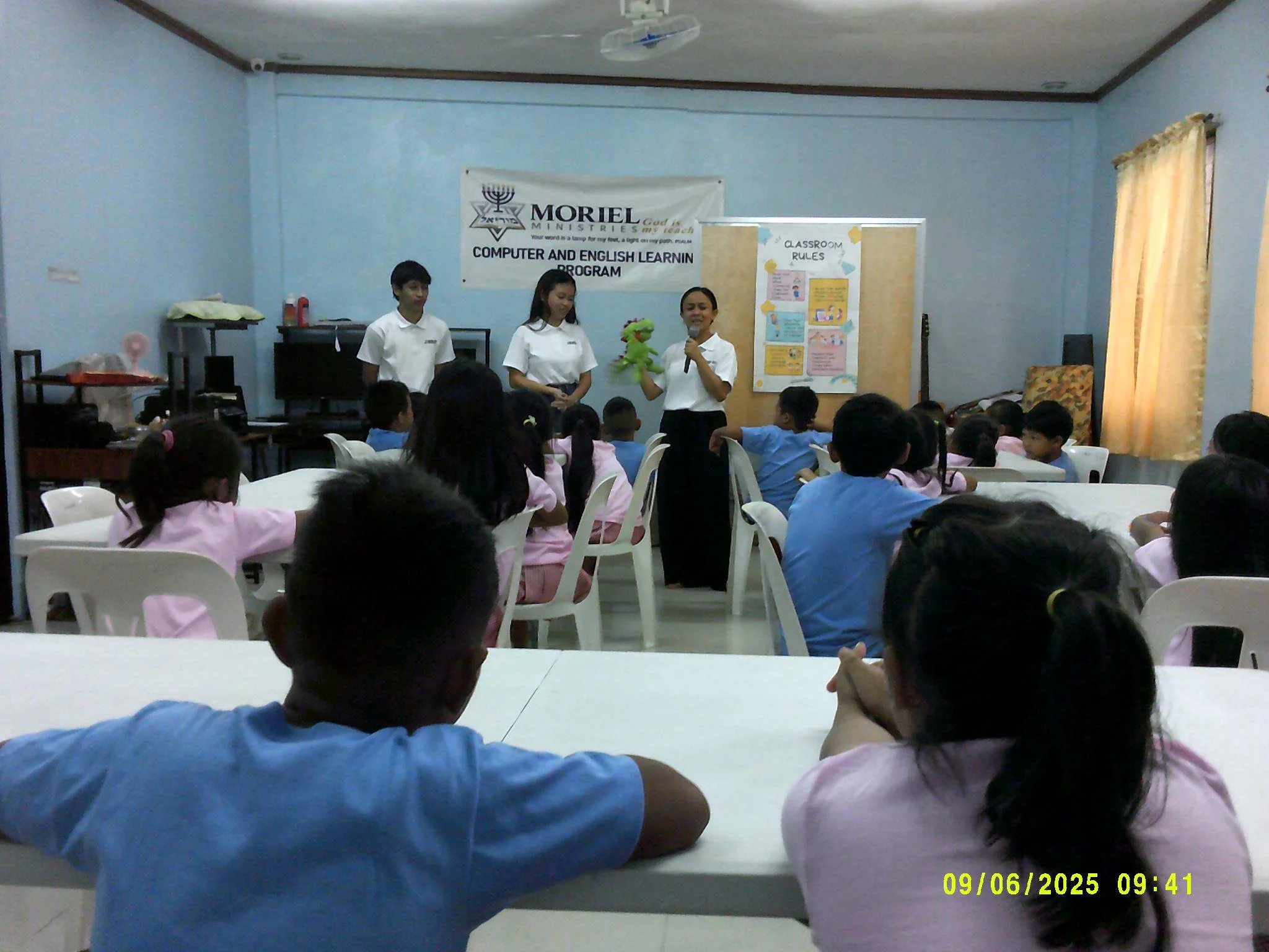 Een klaslokaal met jonge leerlingen die naar een presentatie kijken. Drie volwassenen staan voor de klas, een vrouw houdt een groene knuffel vast en spreekt. Op de achtergrond een wit bord met regels en een banner met de tekst "Morel Ministries".