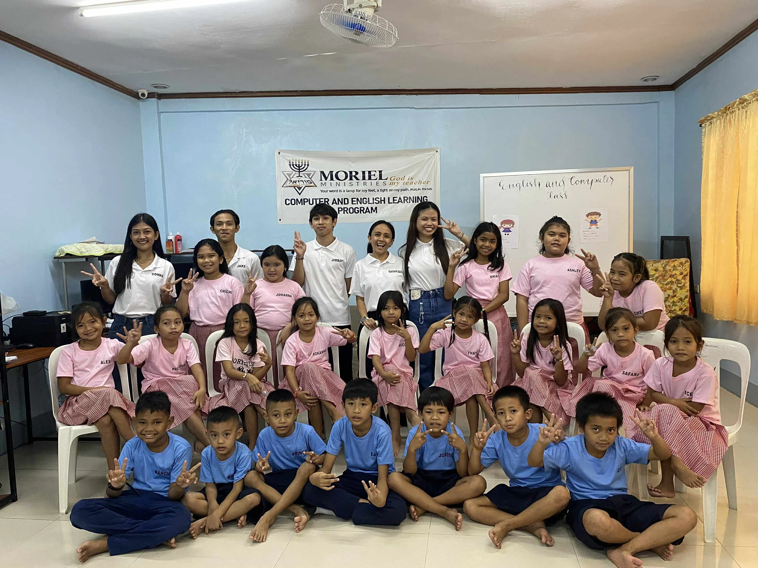 Groep kinderen en leraren poseert in een klaslokaal, sommigen maken vredes- en duim-gebaren.