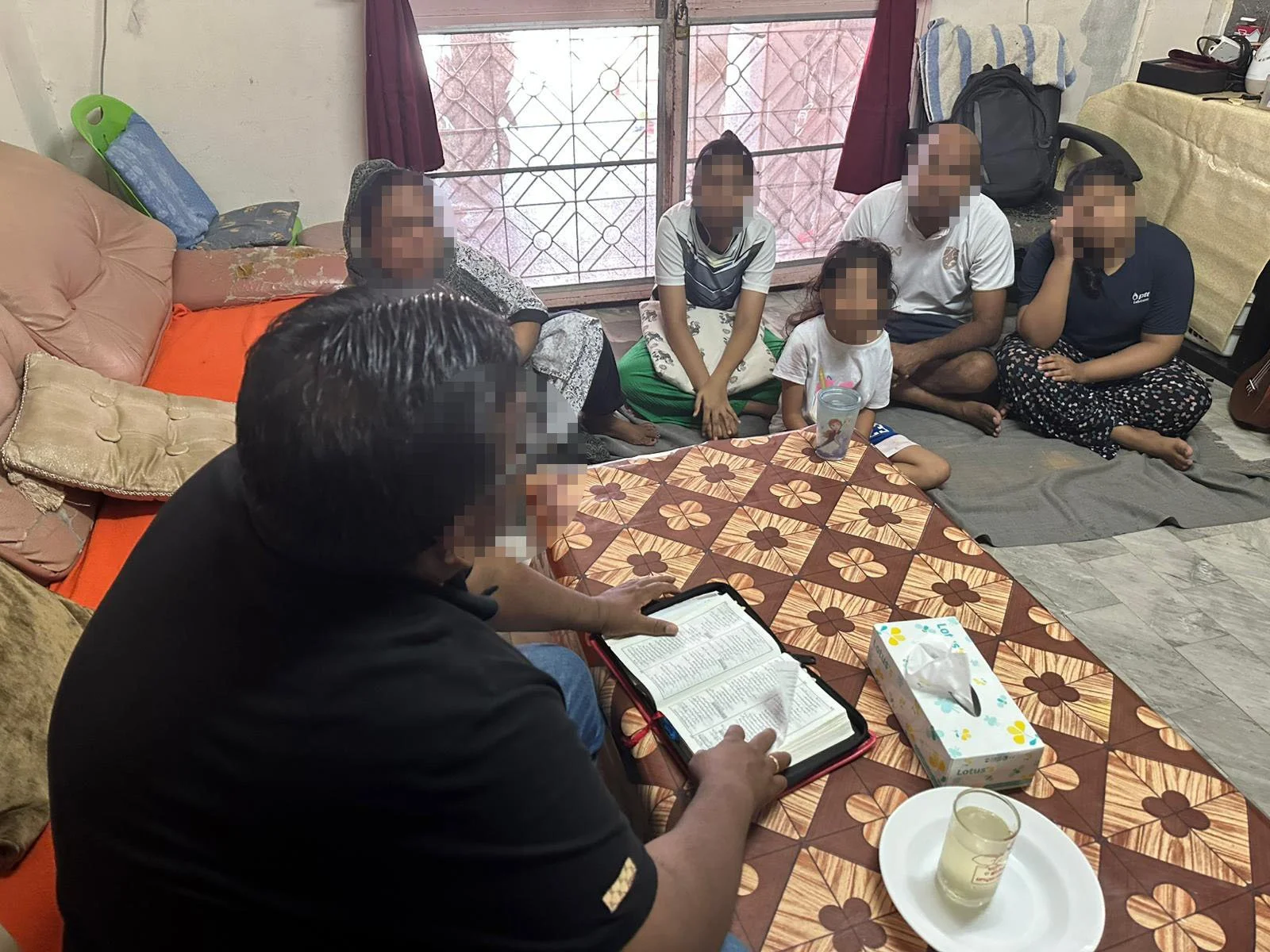 Een binnenruimte met acht mensen die op de vloer van een kamer zitten. Ze luisteren naar een man die een bijbel vasthoudt, die voor een tafel met een tissuedoos, een kaars en een glas drinken staat. De personen maken zich klaar voor een religieuze bijeenkomst of gebedsservice.