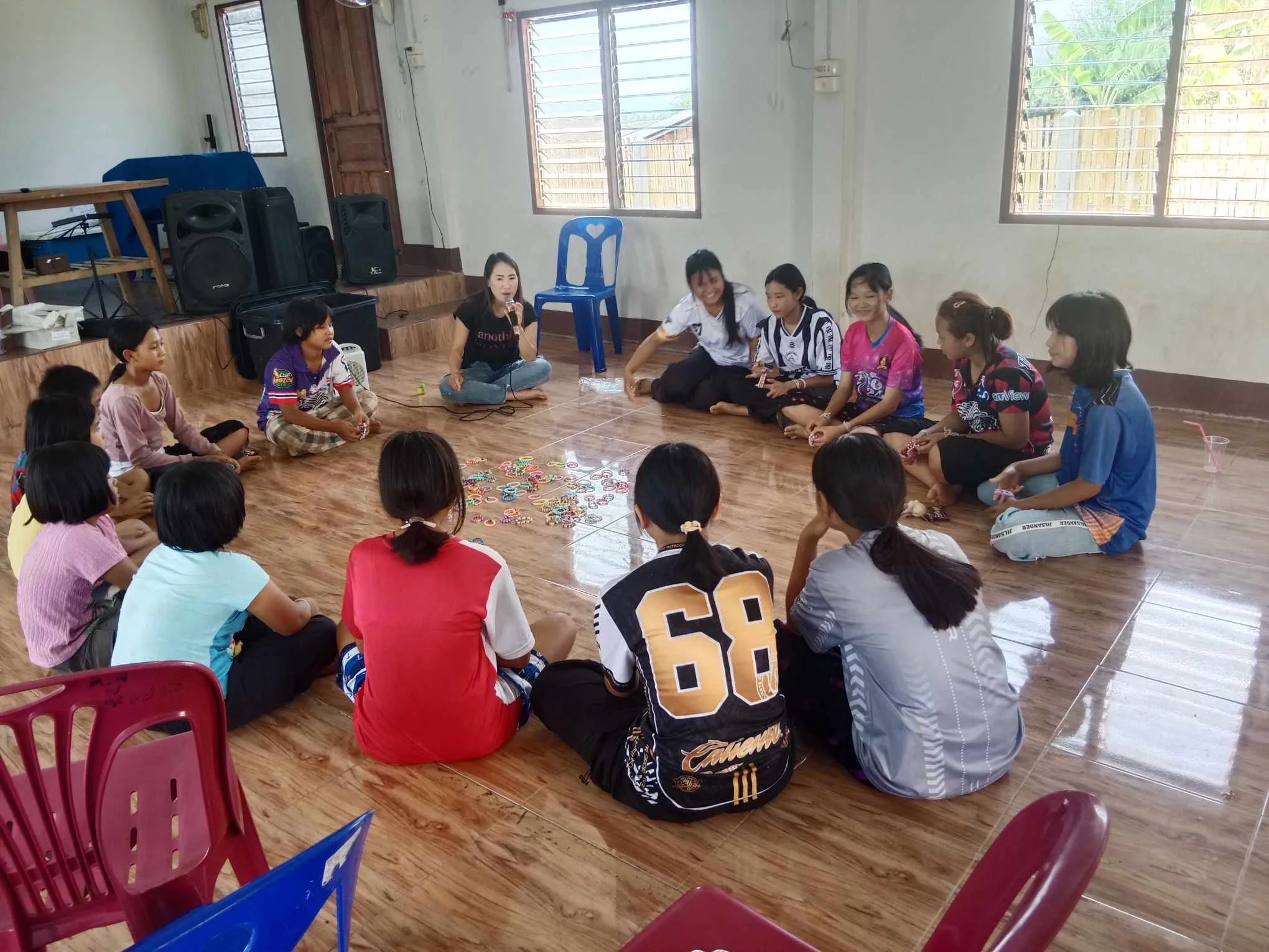 Meisjes en jongens zitten in een cirkel op de houten vloer, met een vrouw die in het midden met een microfoon spreekt, in een kamer met grote ramen en muziekapparatuur op een tafel.