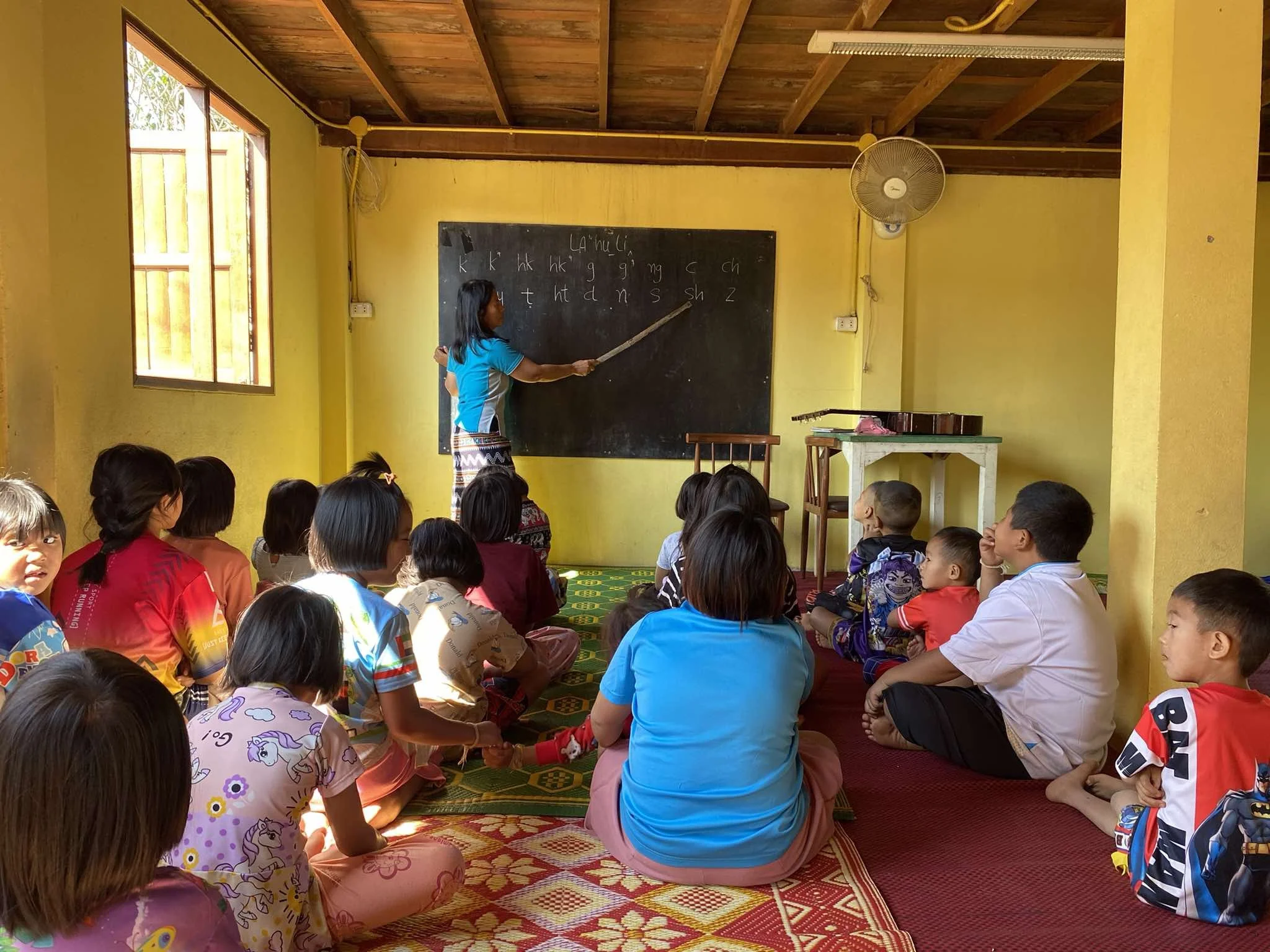 Een leerkracht geeft een les aan een groep jonge kinderen in een klaslokaal. De klas heeft gele muren, een raam dat zonlicht binnenlaat, en een schoolbord met het alfabet. De kinderen zitten gekruist op de vloer, naar de leerkracht kijkend.