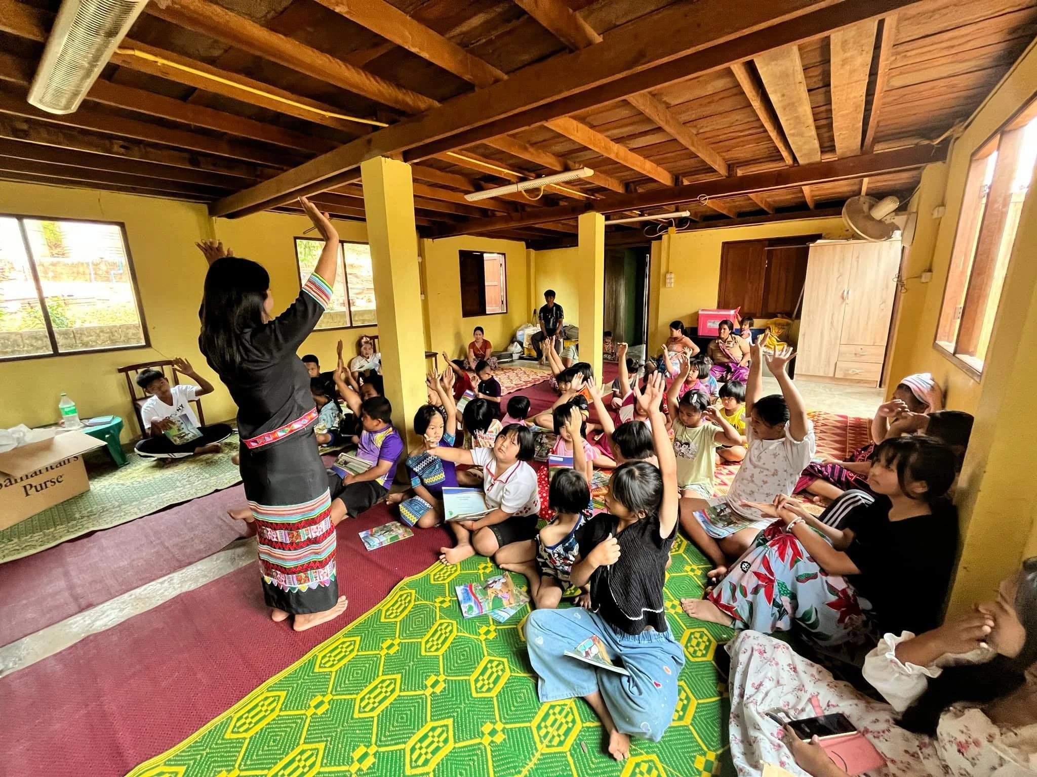 Een groep kinderen zit op de vloer van een kamer, allemaal met hun handen omhoog, terwijl een vrouw vooraan in traditionele kleding ze onderwijst of instrueert. De kamer heeft gele muren en houten plafonds, met ramen die natuurlijk licht binnenlaten.