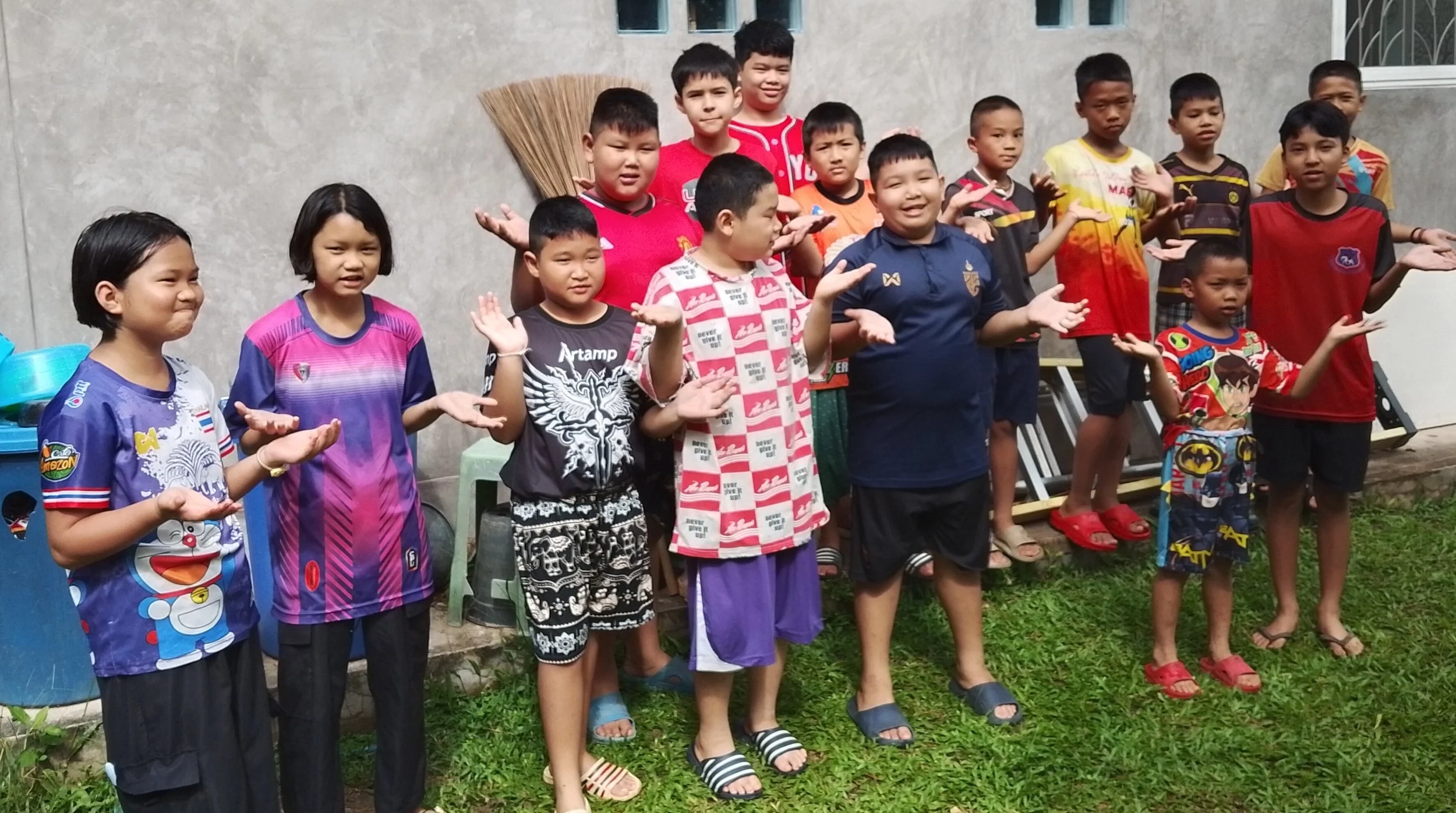 Groep kinderen staan samen buiten, met armen uitgestoken. Sommigen glimlachen, anderen kijken serieus. Op de achtergrond een muur en een raam.