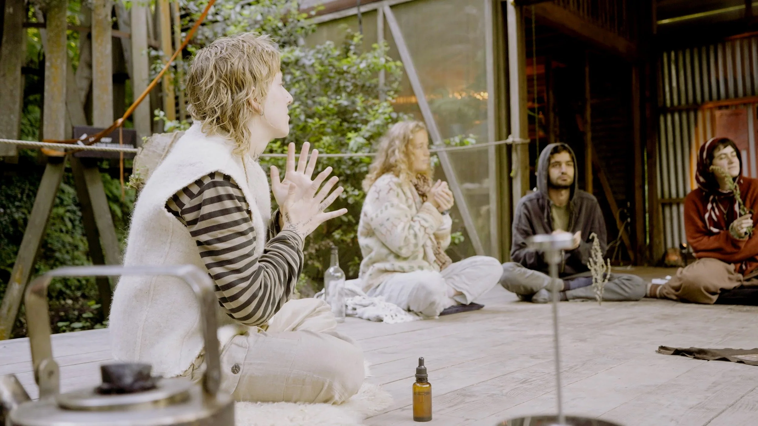 Four people sitting cross-legged on a wooden floor outdoors, in a circle, with greenery and a wooden structure in the background, participating in a meditation or prayer session.