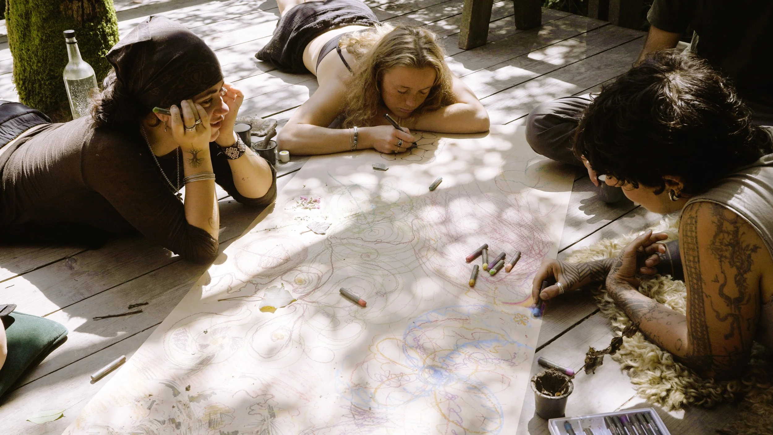 Three women are lying on a wooden porch, drawing and coloring on a large sheet of paper spread out on the floor. The woman on the left has a headscarf and tattoos, the woman in the middle has curly hair and is focused on her drawing, and the woman on the right has dark curly hair, tattoos, and is using colored chalks.