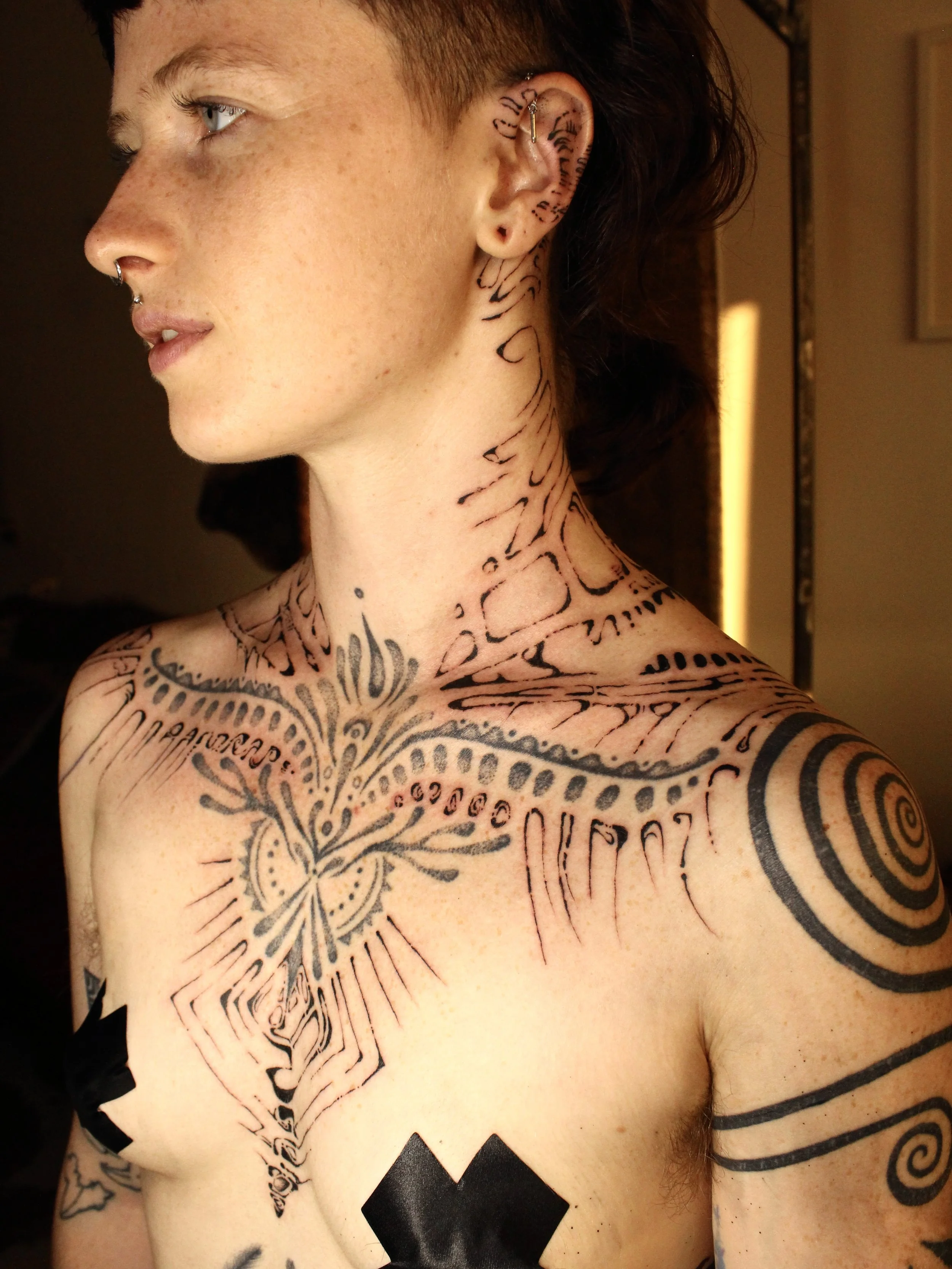 A young person with short hair, multiple facial piercings, and extensive black tattoos on their neck, chest, shoulder, and arm, including geometric patterns, spiral designs, and tribal motifs.