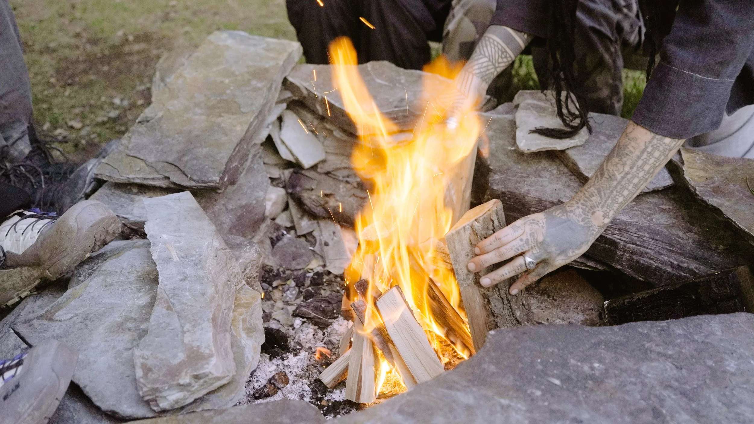 Person with tattooed arms building a campfire with wooden logs surrounded by large stones.