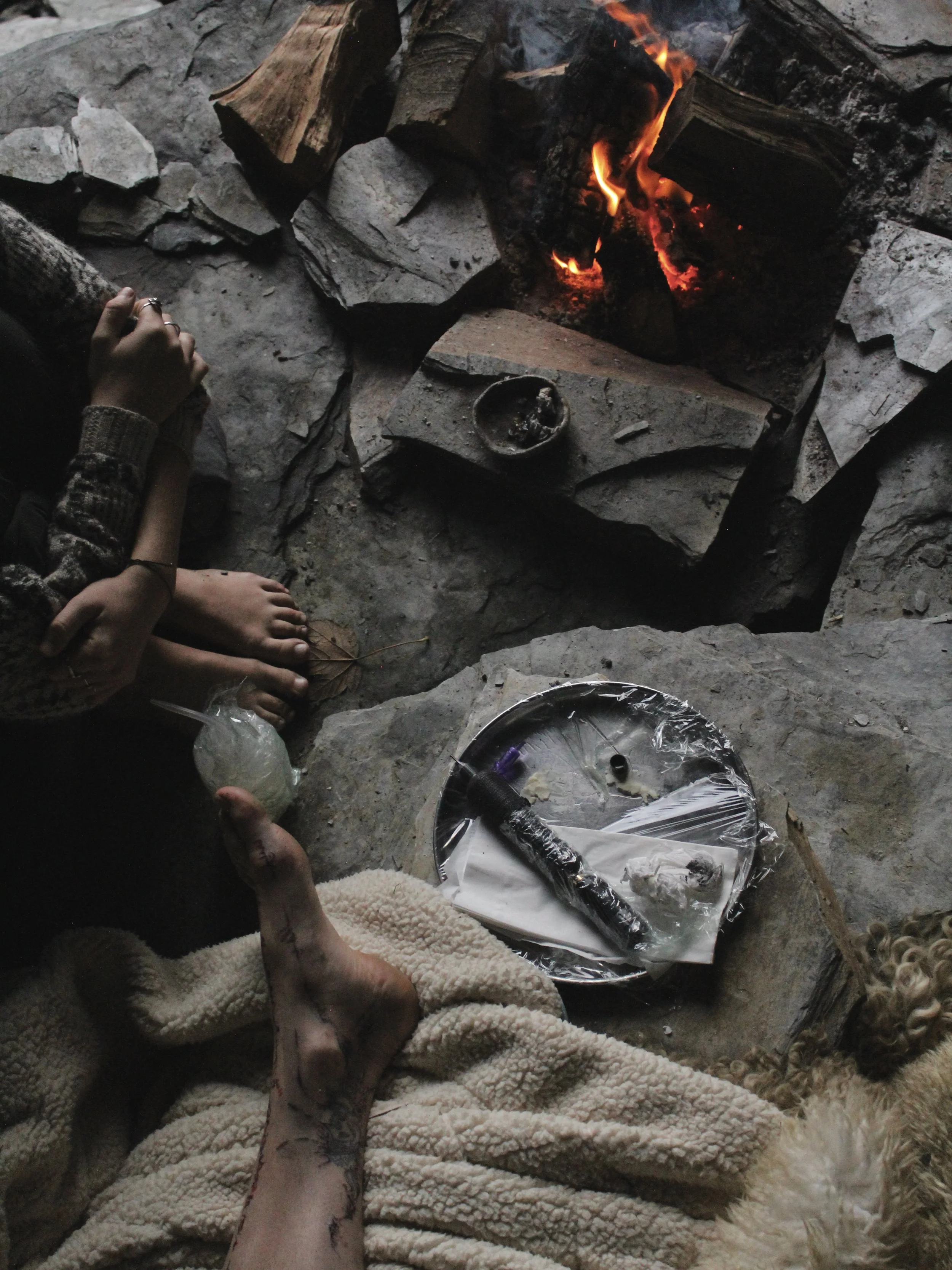 A cozy indoor scene with a small fire burning on a stone hearth, surrounded by rocks and logs, with a person's bare foot and hand visible, a tray with indicating evidence, and a dog lying nearby.