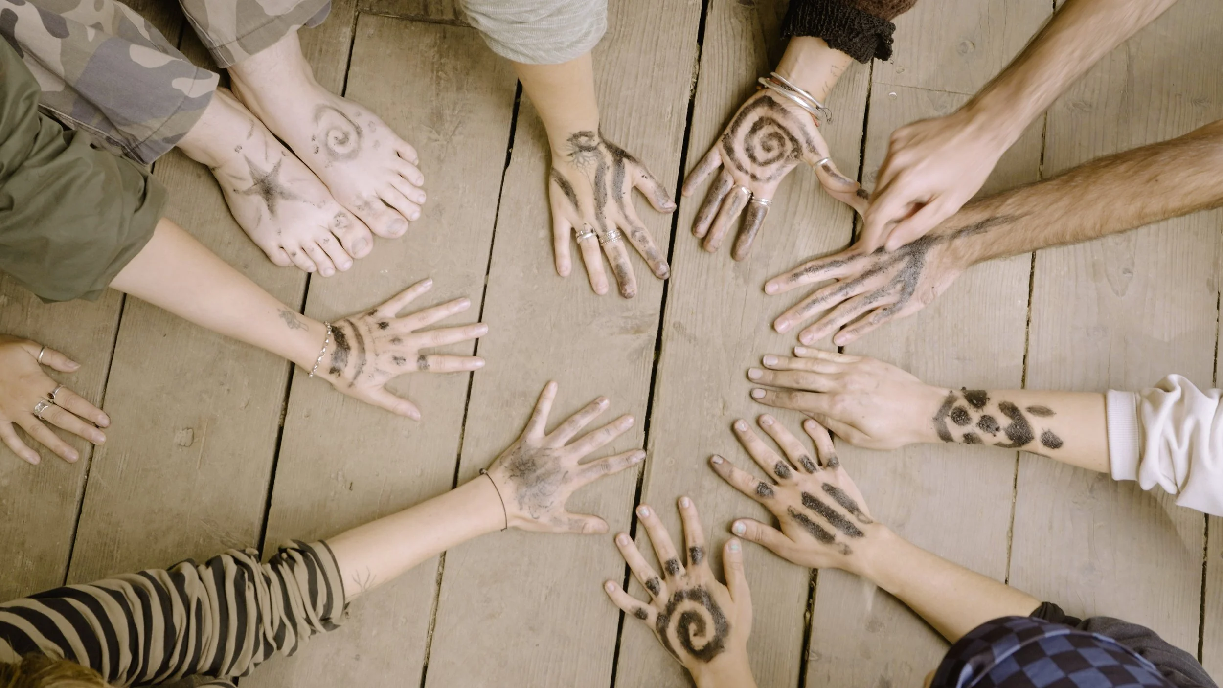 A group of people in a circle showing their hands and feet on a wooden floor, with each hand and foot decorated with black ink drawings and tattoos.