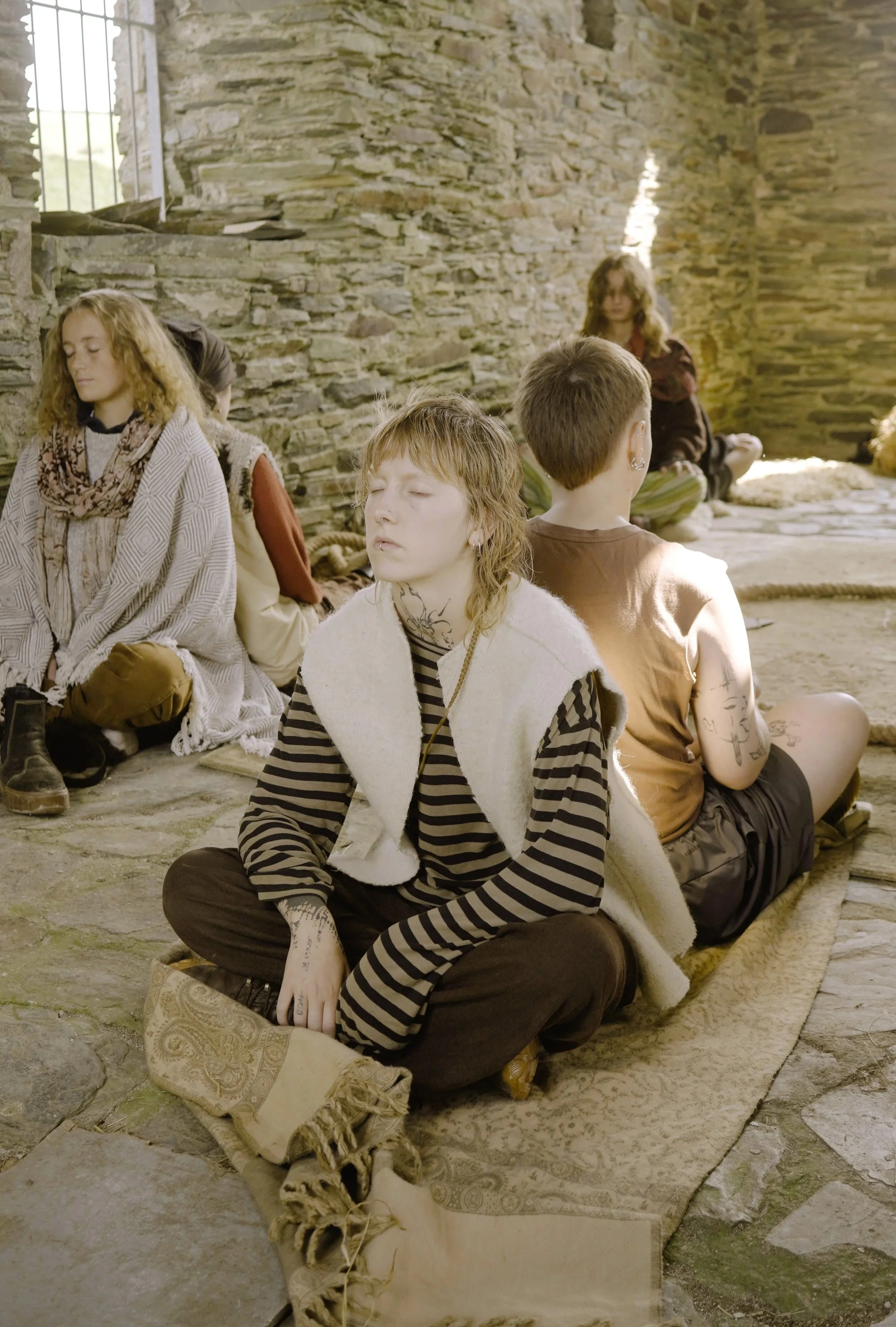 Group of young people sitting cross-legged on a carpet in a stone-walled room, with some appearing to meditate or relax with closed eyes.
