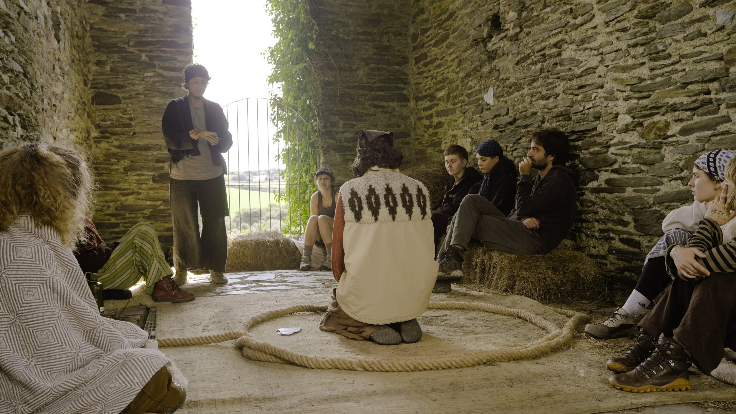 A group of people sitting inside a stone-walled shelter with an open gate, listening to a person standing and speaking. The setting appears rustic, with hay bales and a natural background visible through the open gate.