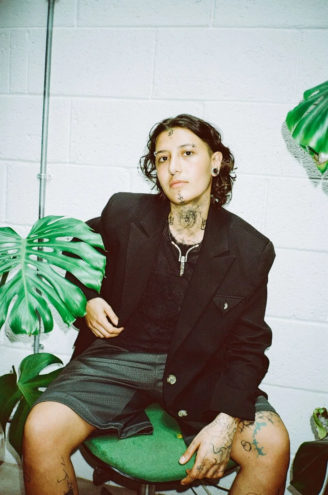 Person sitting on a green office chair, wearing a black blazer and shorts, with tattoos on their arms, hands, face, and neck, in front of a white cinder block wall, surrounded by large green indoor plants.