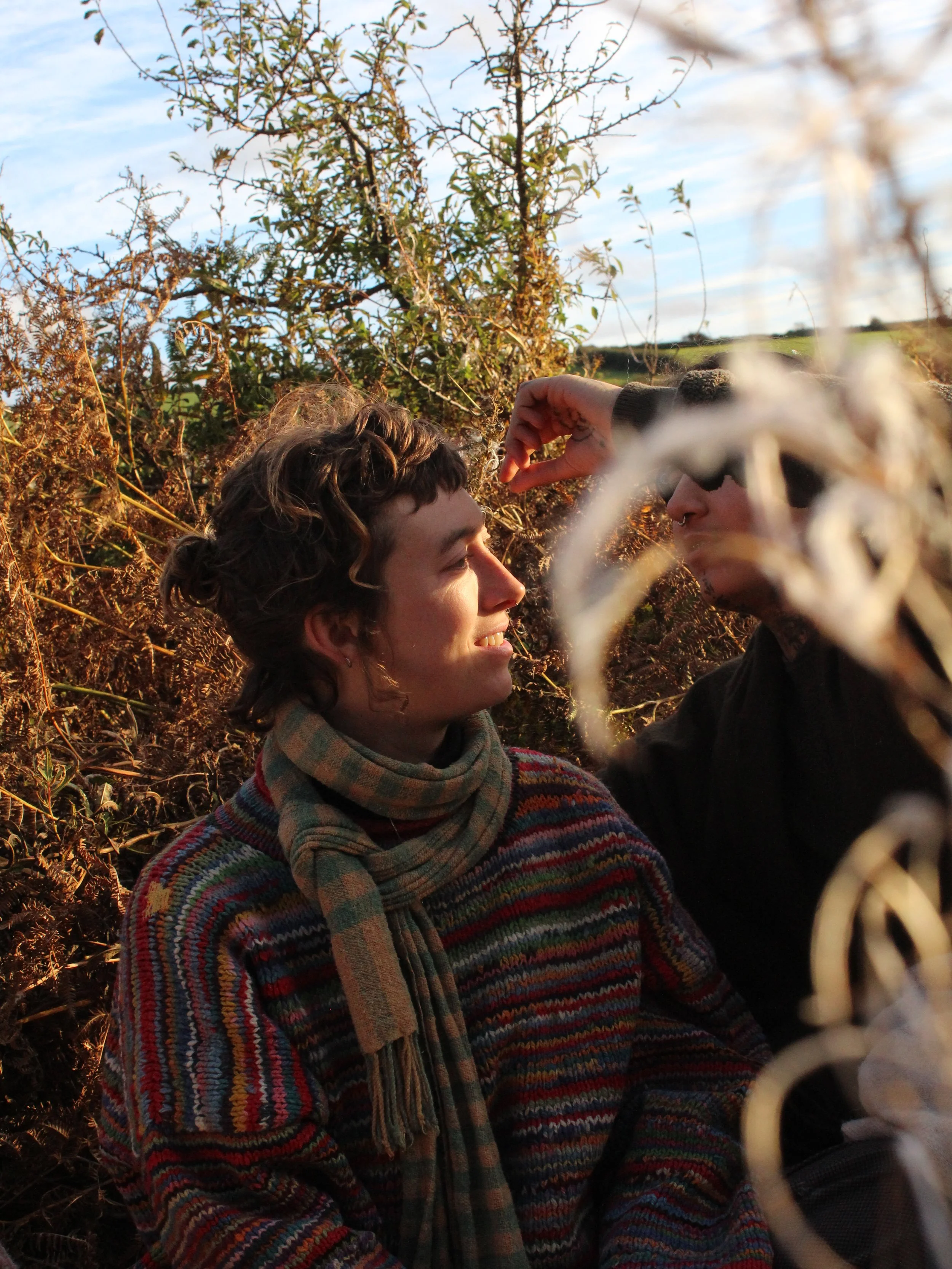 Two individuals with short, dark hair, sitting outdoors in a natural, wooded area, one with a scarf, smiling, while the other is adjusting sunglasses, surrounded by brown dried plants and a clear sky.