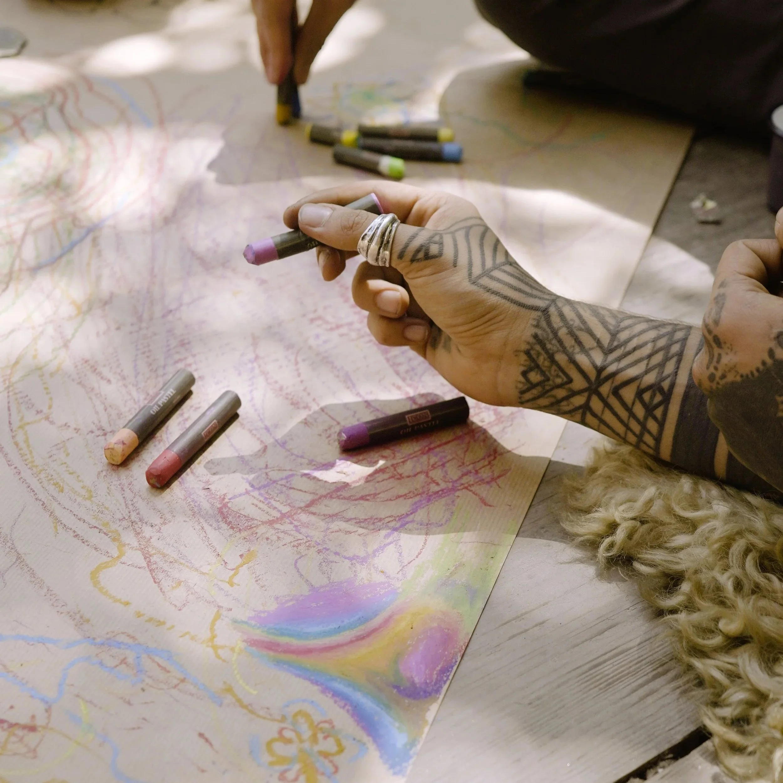 A person with tattooed hands drawing colorful lines and patterns on a large sheet of paper with pastel sticks, sitting on a wooden floor.