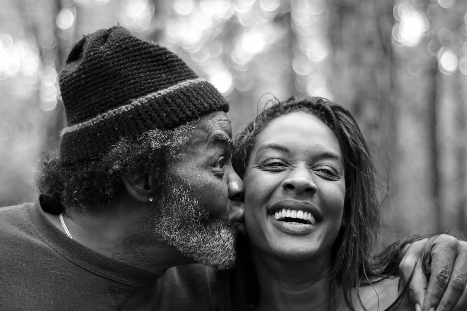 A man with glasses, a beard, and a beanie hat is kissing a smiling woman on the cheek, both outdoors in a forest.