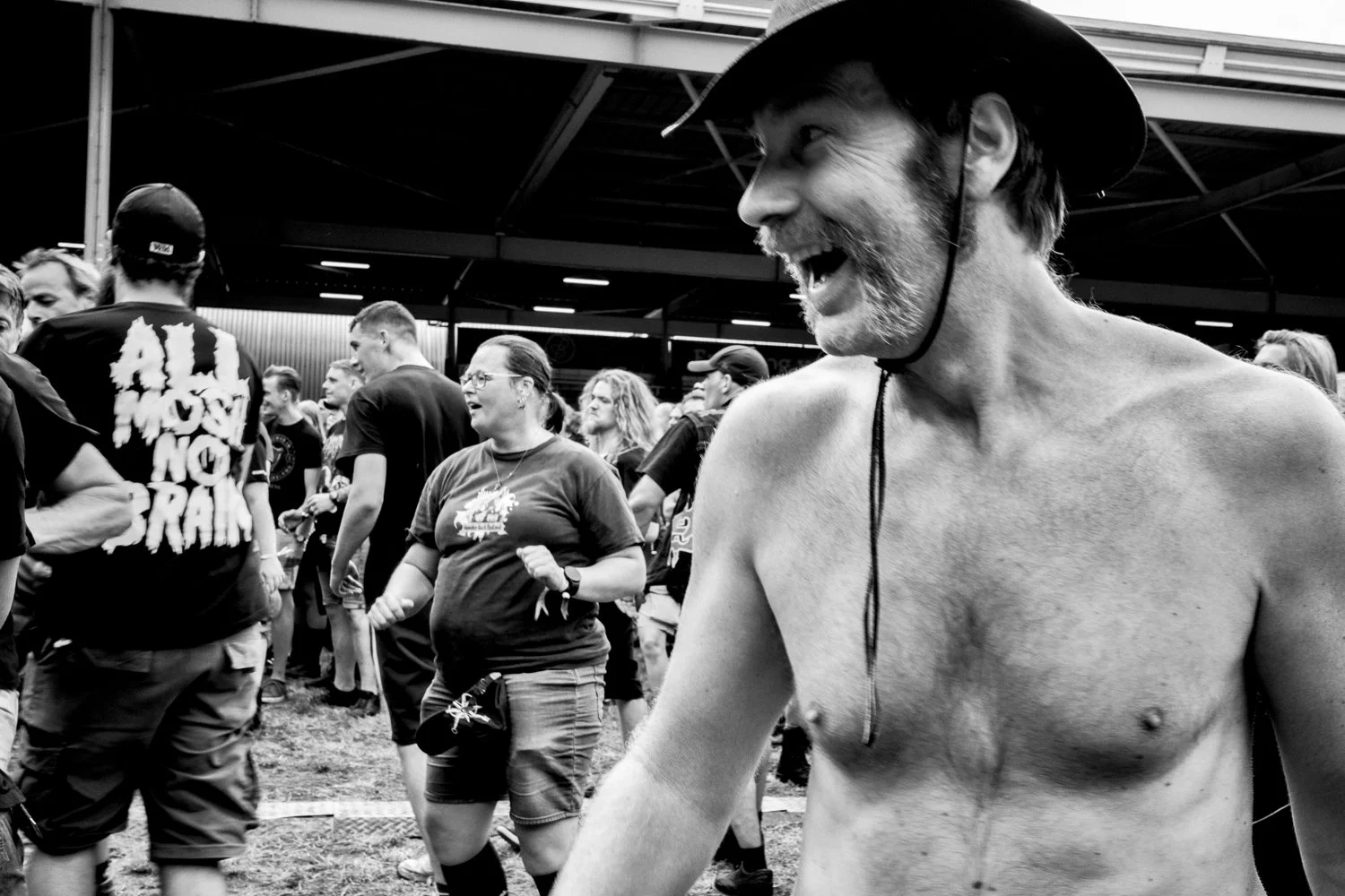 A shirtless man with a beard and wide-brimmed hat smiling amid a crowd at an outdoor event, with people dancing and socializing in the background.