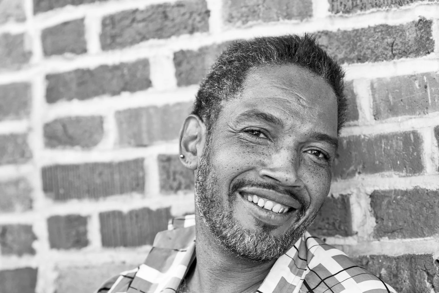 A smiling man with a beard and earrings standing against a brick wall.