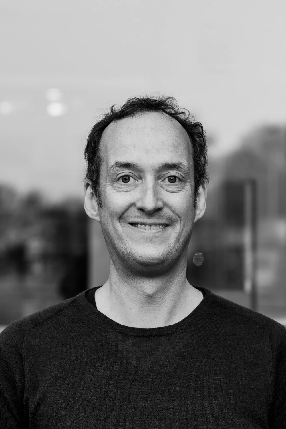 Black and white portrait of a man with short dark hair, wearing a dark sweater, standing outdoors with blurred background of buildings and sky.