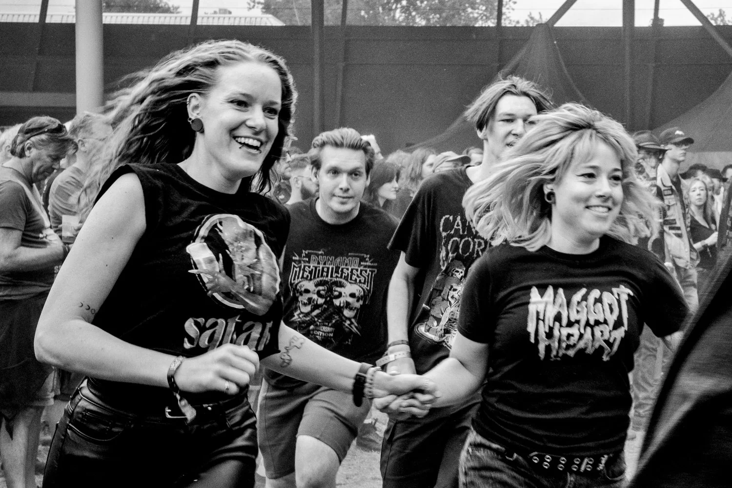 Two young women with light skin and wearing black band t-shirts, smiling and holding hands, at an outdoor music festival with other people in the background.