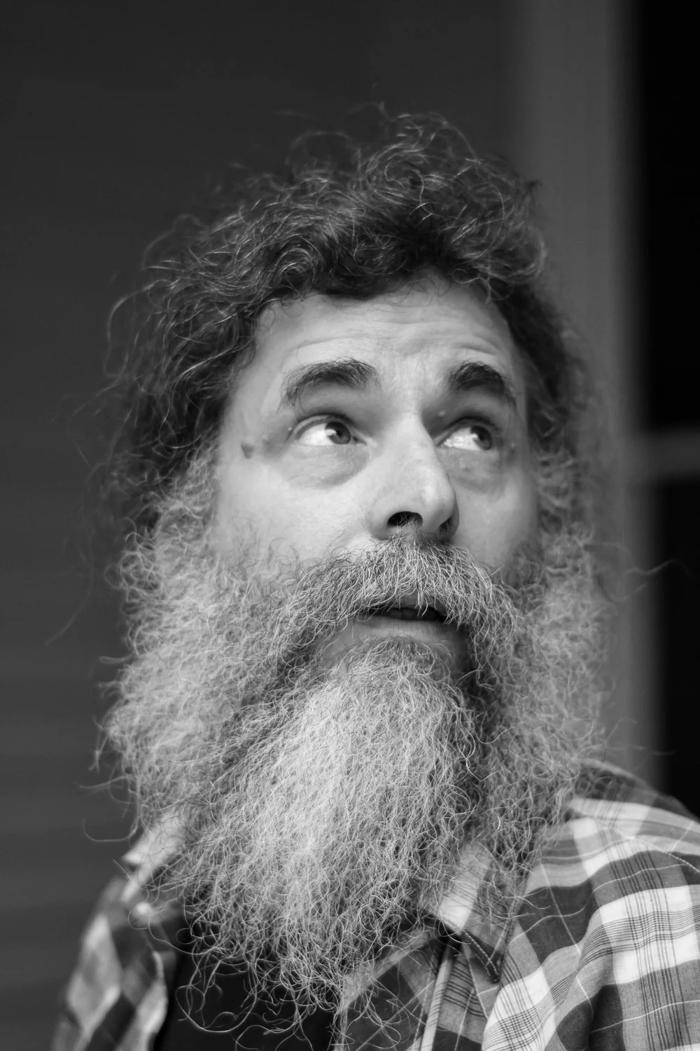 Black and white close-up portrait of a man with curly hair and a full beard, looking upward and to the side, wearing a plaid shirt.