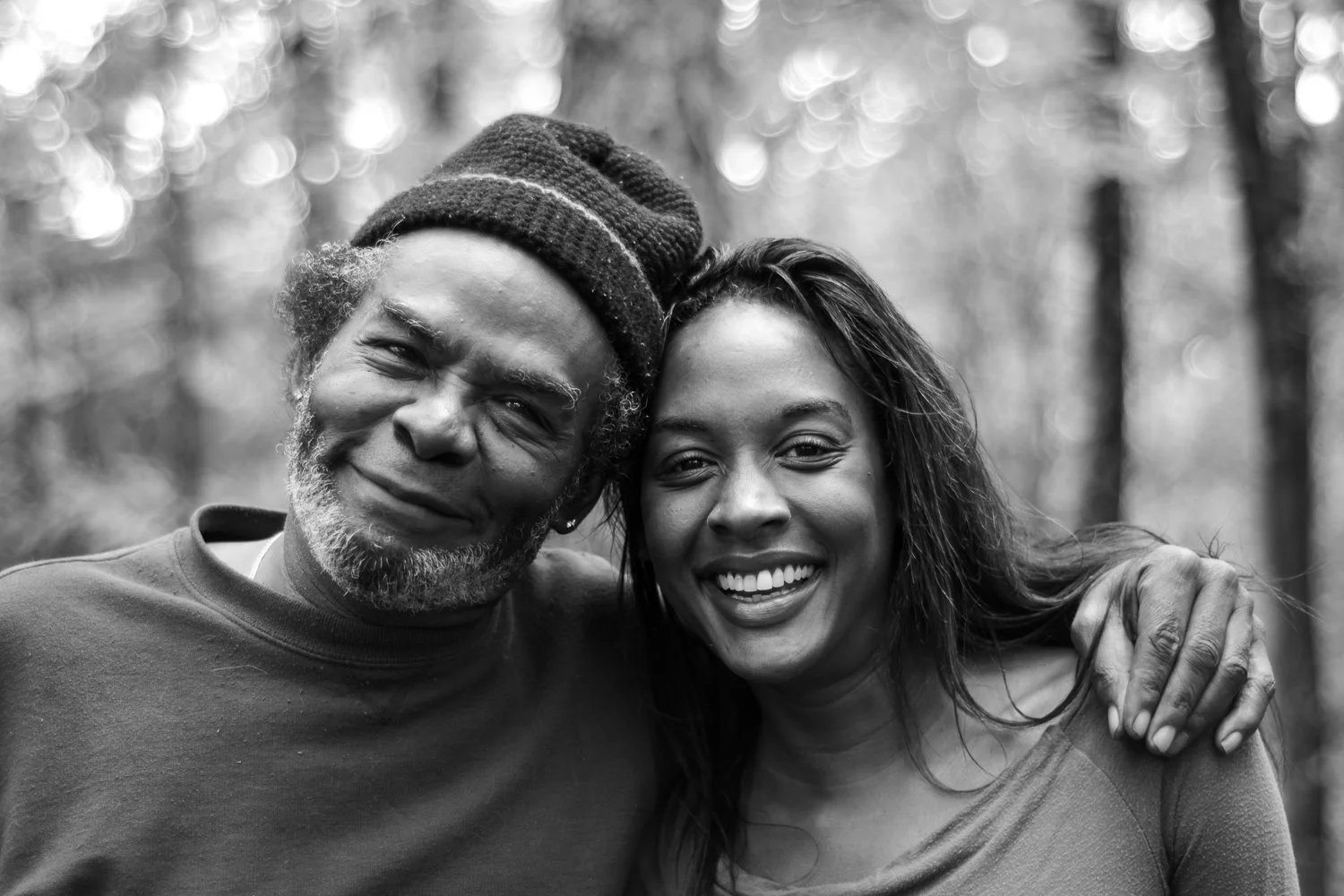 A happy older man and a young woman smiling together outdoors in a forest, with the man wearing a beanie and the woman having long wavy hair.