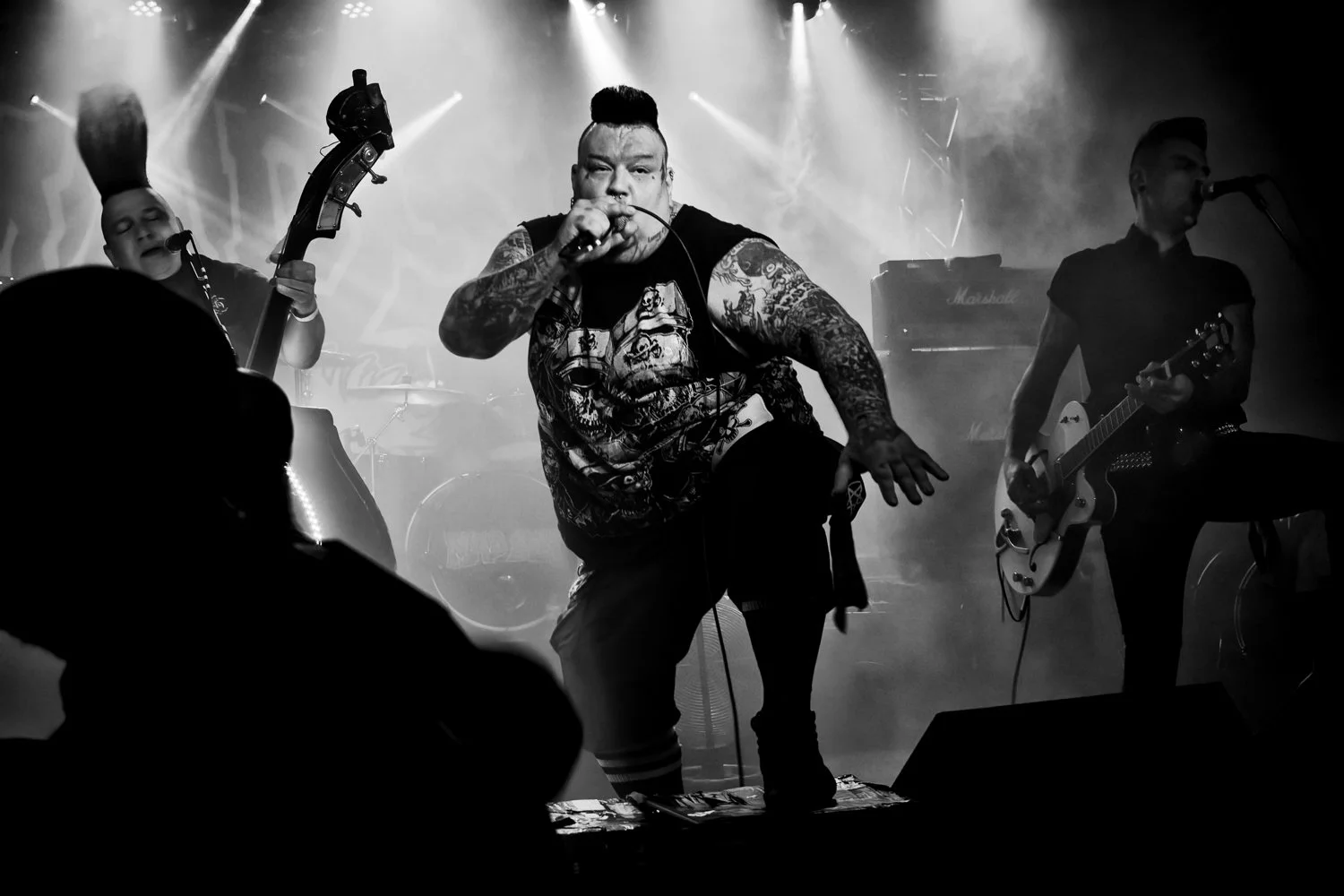 A black-and-white photo of a band performing on stage, featuring a lead singer with tattoos, a mohawk hairstyle, and a graphic t-shirt, accompanied by two other musicians playing guitar and bass.