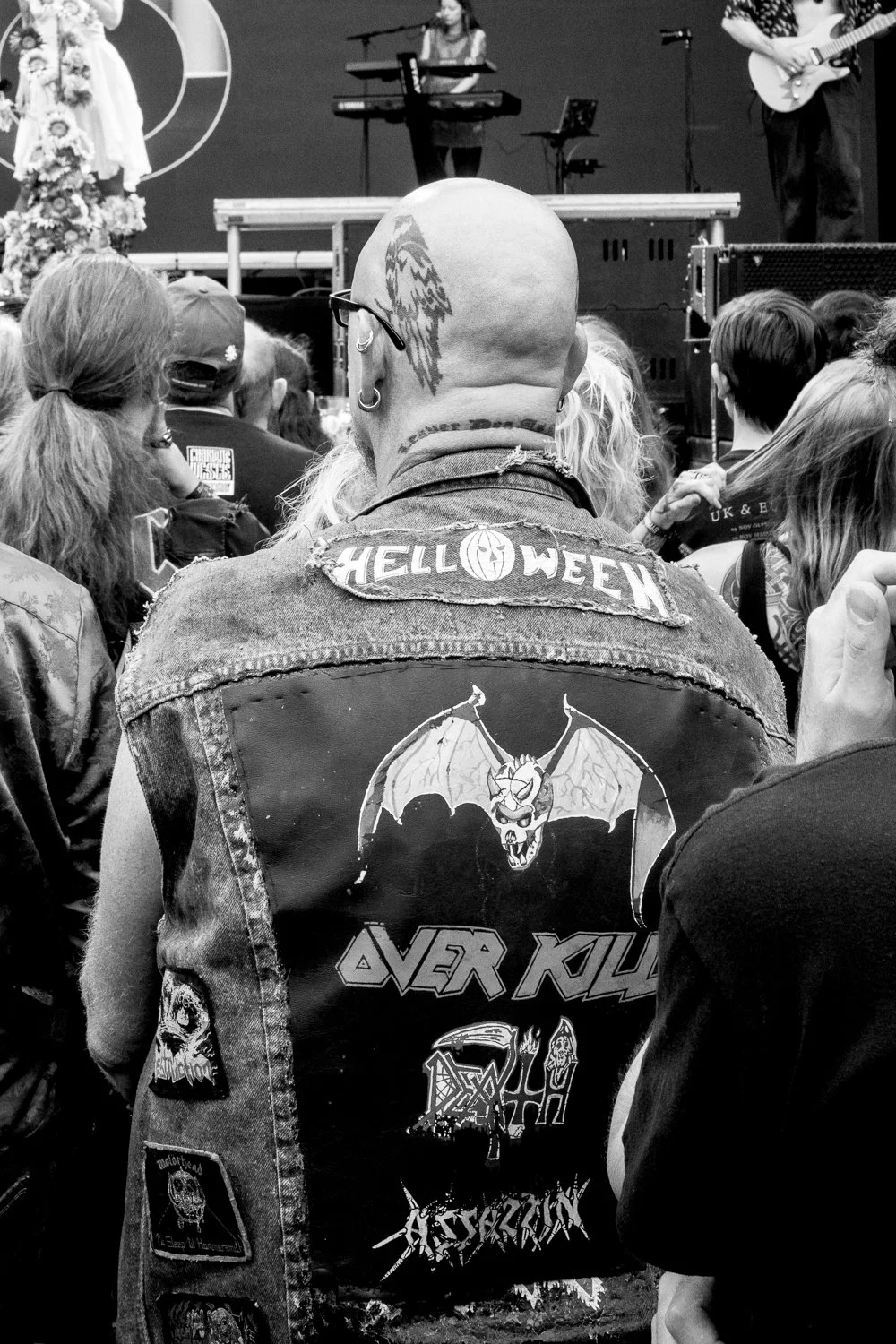 A person with a shaved head and tattoos on their neck and head, wearing a denim jacket with patches and graphic designs, at a concert with a crowd of people and a stage with musicians in the background.