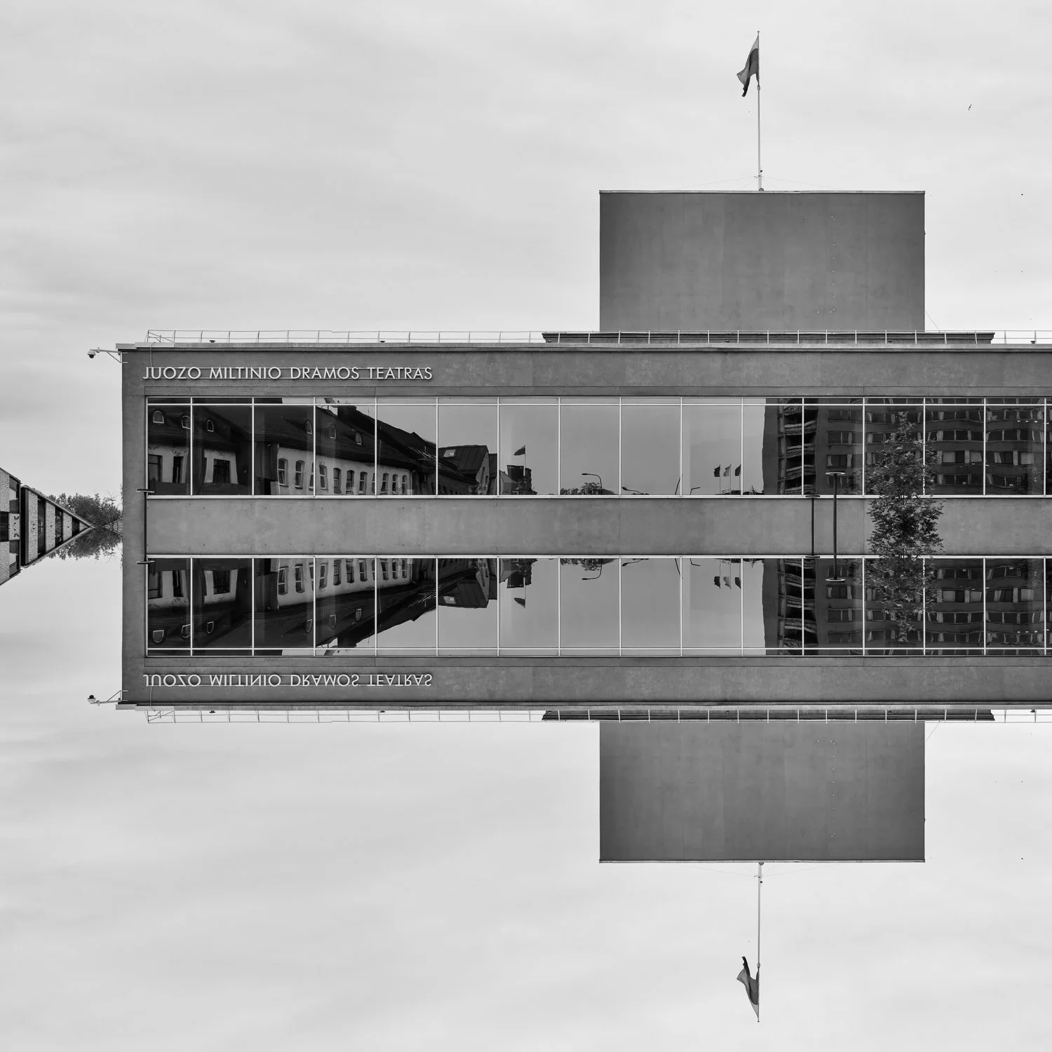 Black and white photo of a modern building with glass windows, reflecting surrounding buildings, with a flag on top and the sign 'JUOZO MILTINIO DRAMOS TEATRAS'. The photo is upside down, creating a mirror effect.